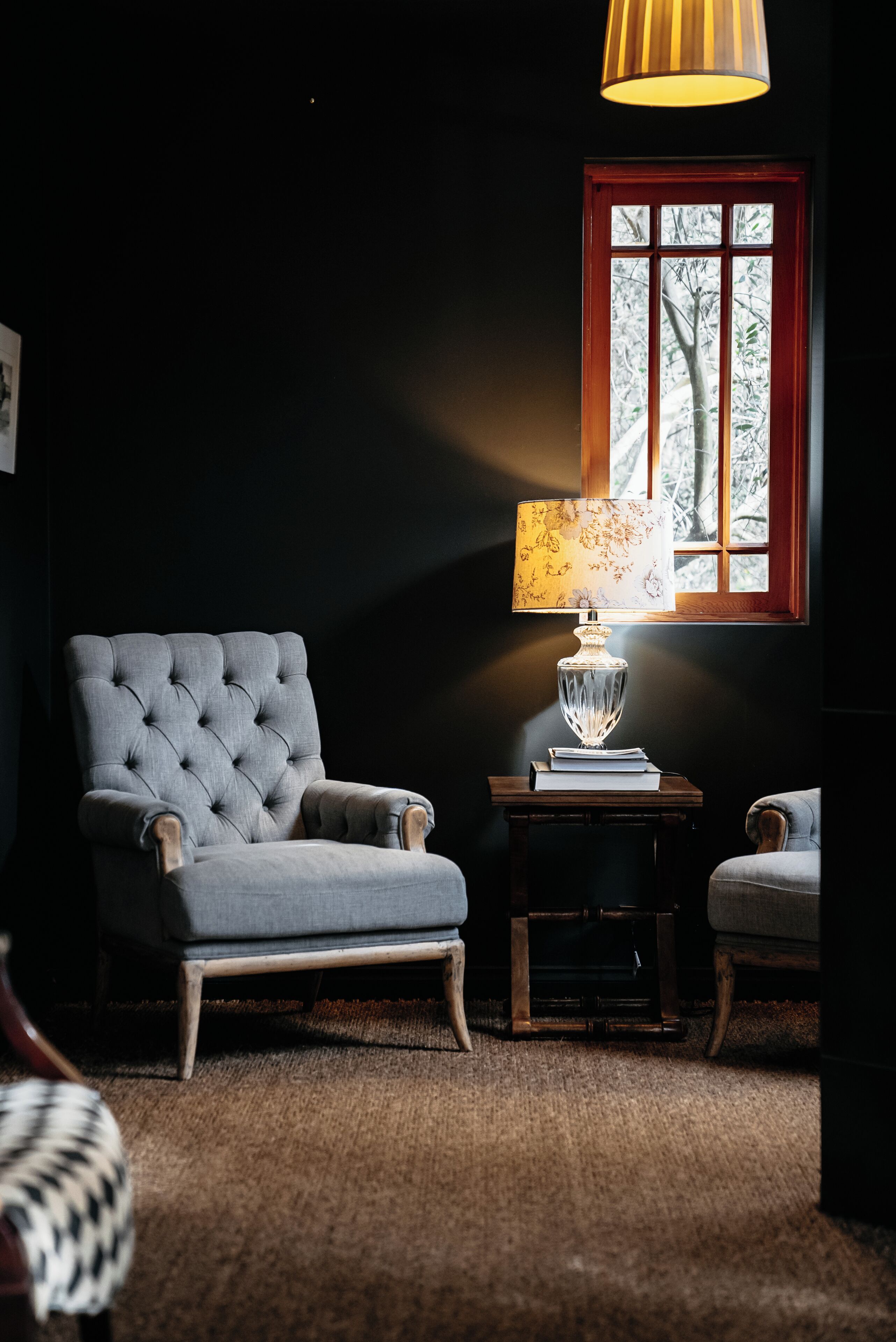 Una habitación con un sillón tapizado, una mesita con lámpara y una ventana con vistas a árboles.