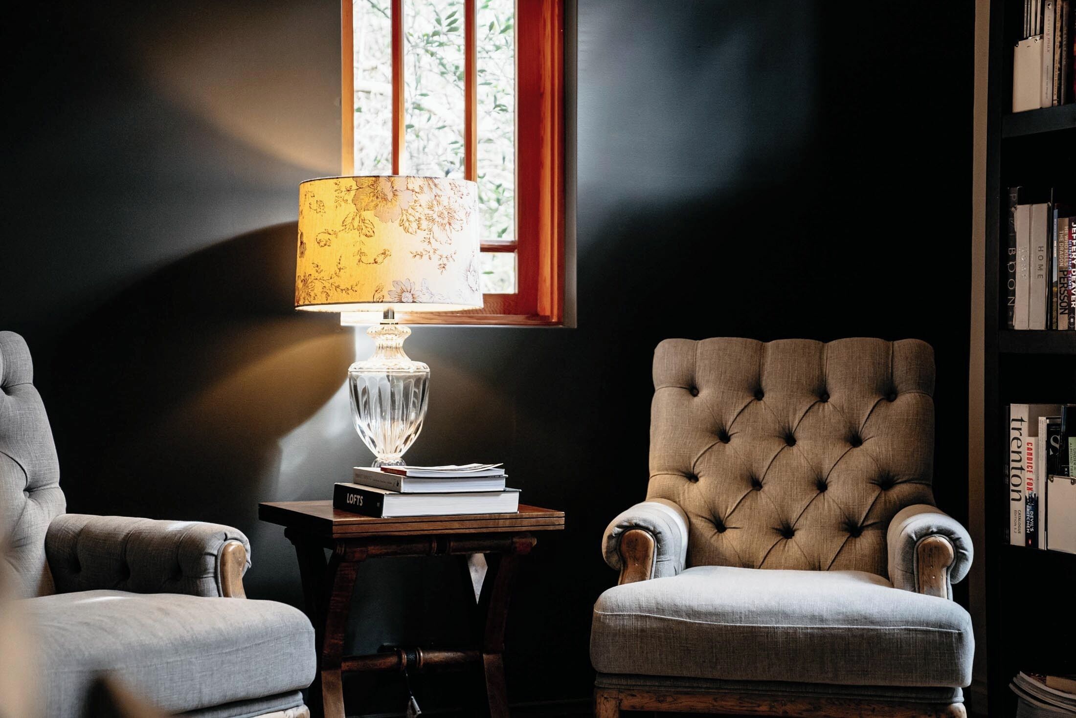 A room with a dark wall, a window with vertical blinds, a lamp with a floral lampshade, a tufted armchair, a smaller armchair, and a bookshelf with books.