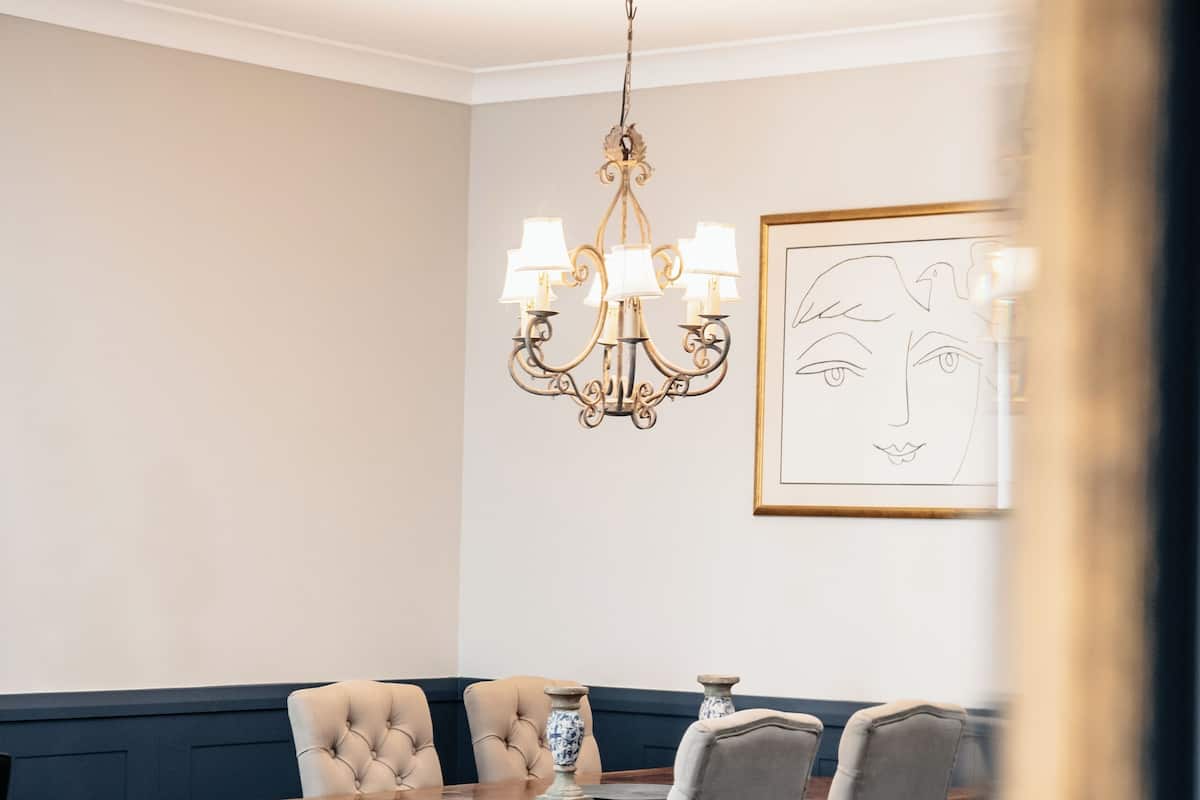 A dining area with a wooden table, cushioned chairs, a chandelier, and a framed artwork on the wall.