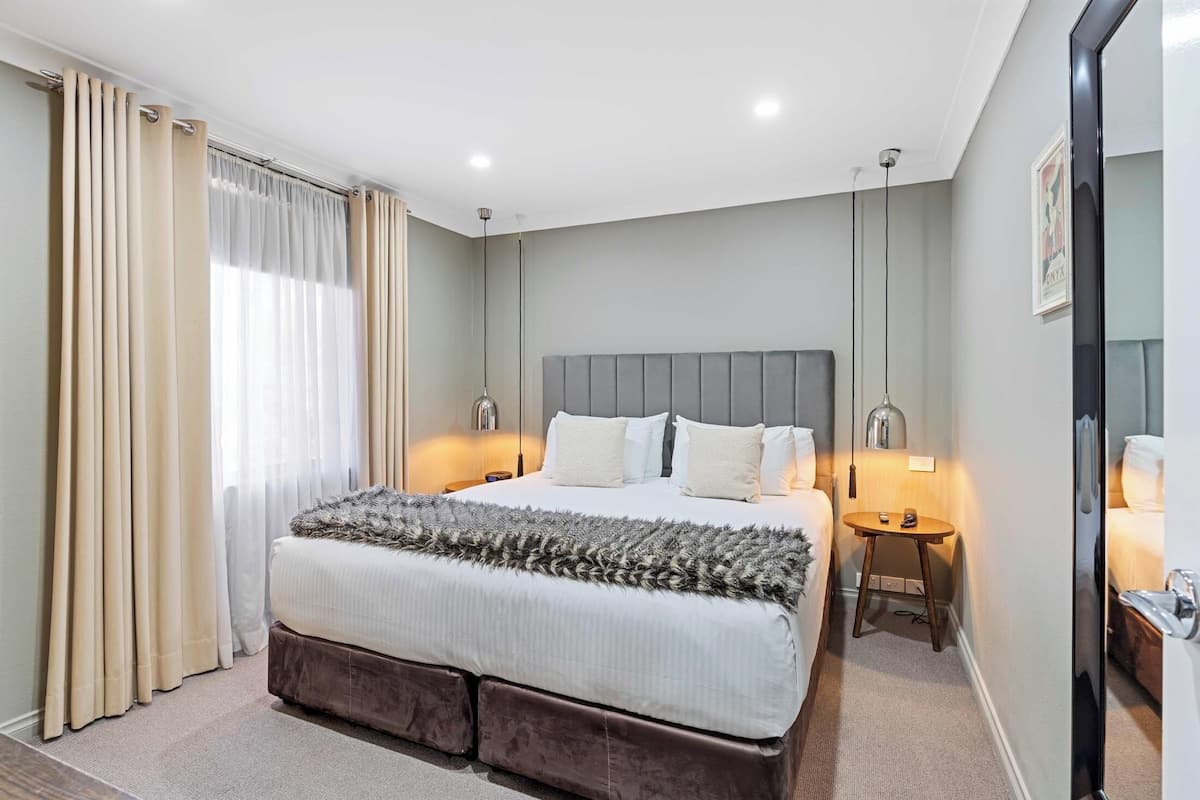 A modern hotel room with a large bed, two bedside tables, a bedside lamp, a mirror, and a window with curtains.