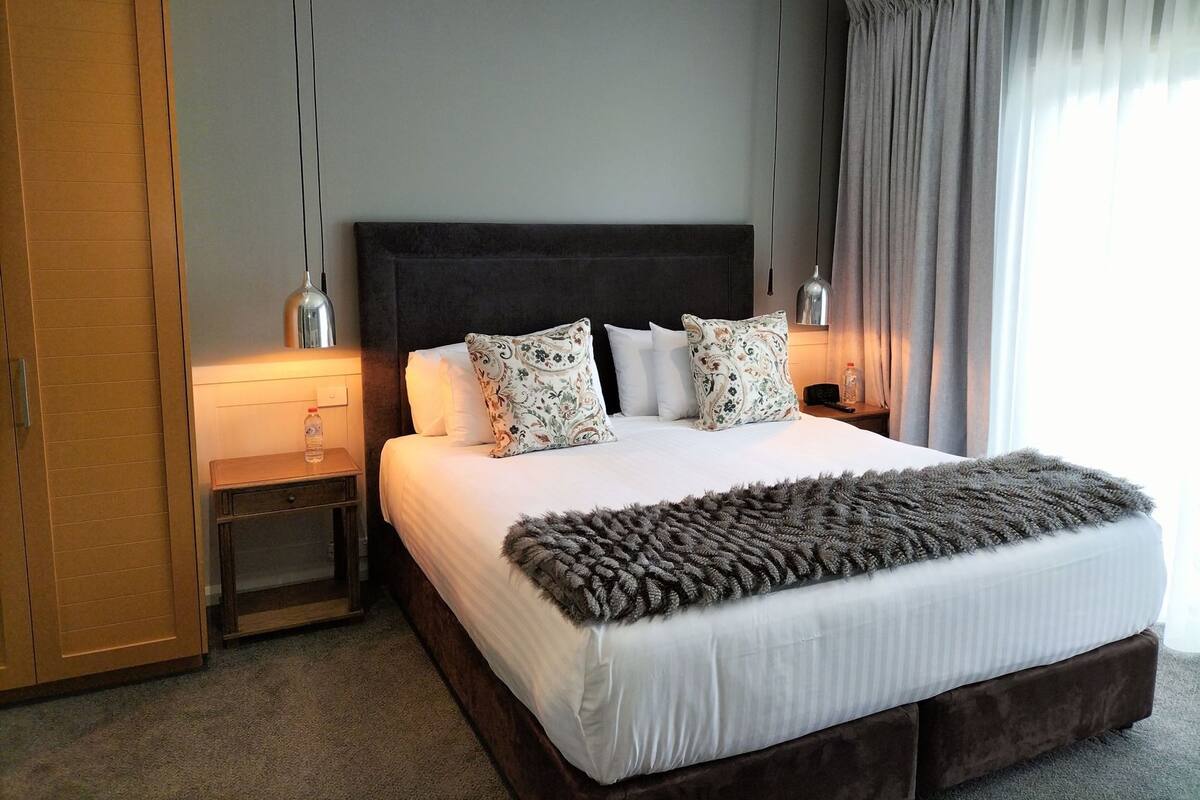 A hotel room with a large bed, two bedside tables, a wardrobe, and a window with curtains.