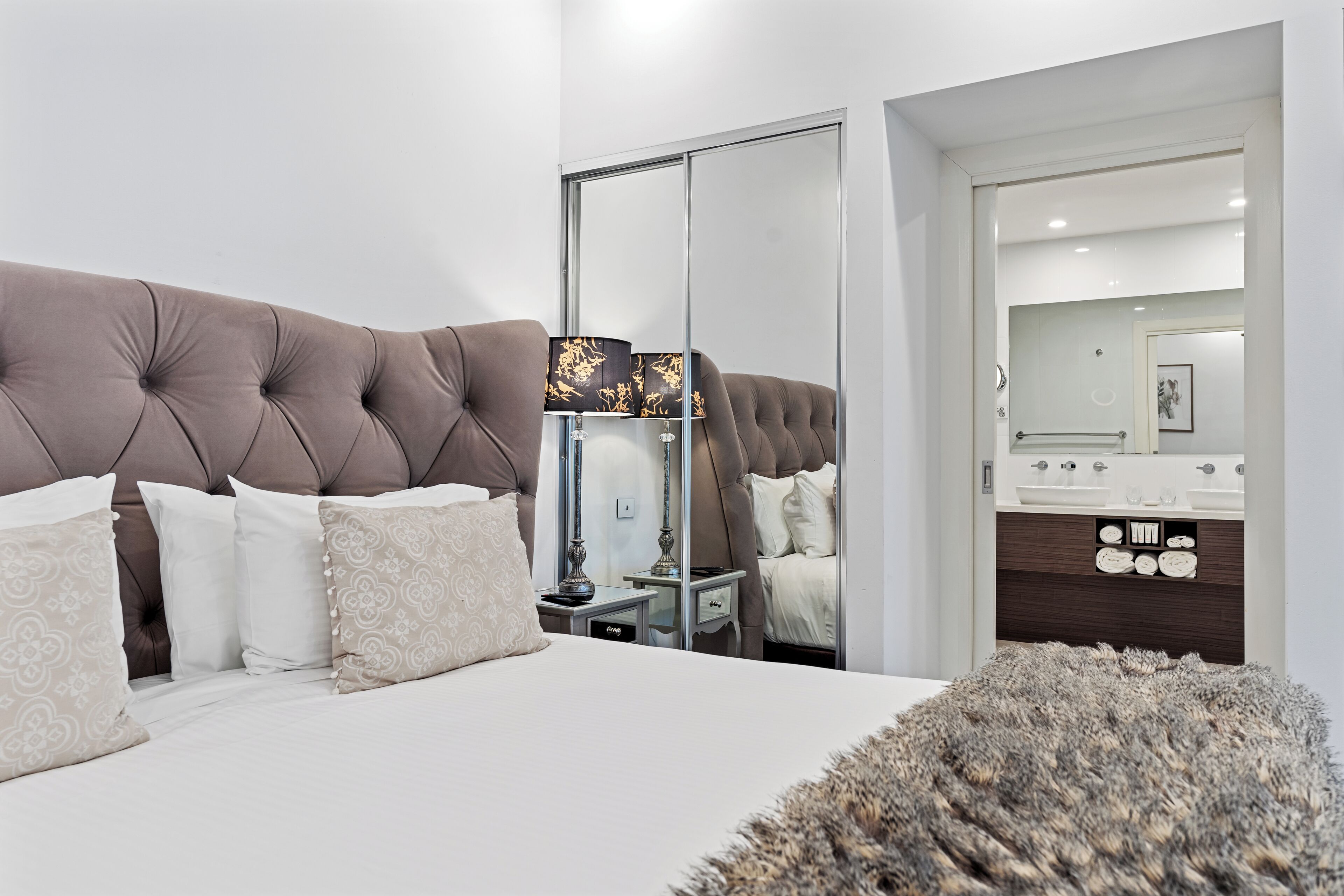 A modern bedroom with a tufted headboard, a large mirror, a bedside table with a lamp, and a fluffy rug.