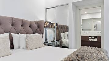 A modern bedroom with a tufted headboard, a large mirror, a bedside table with a lamp, and a fluffy rug.