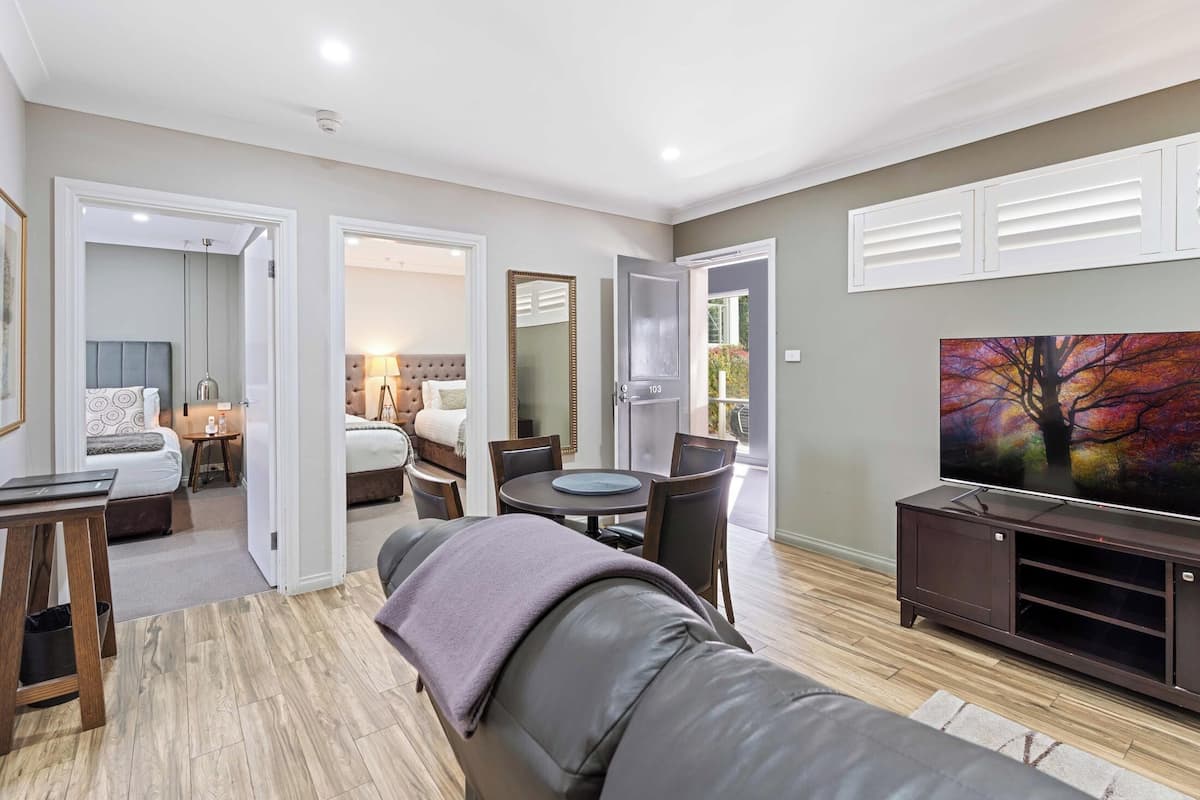 A modern living room with a leather sofa, a dining table with chairs, a flat-screen TV, and a painting on the wall.