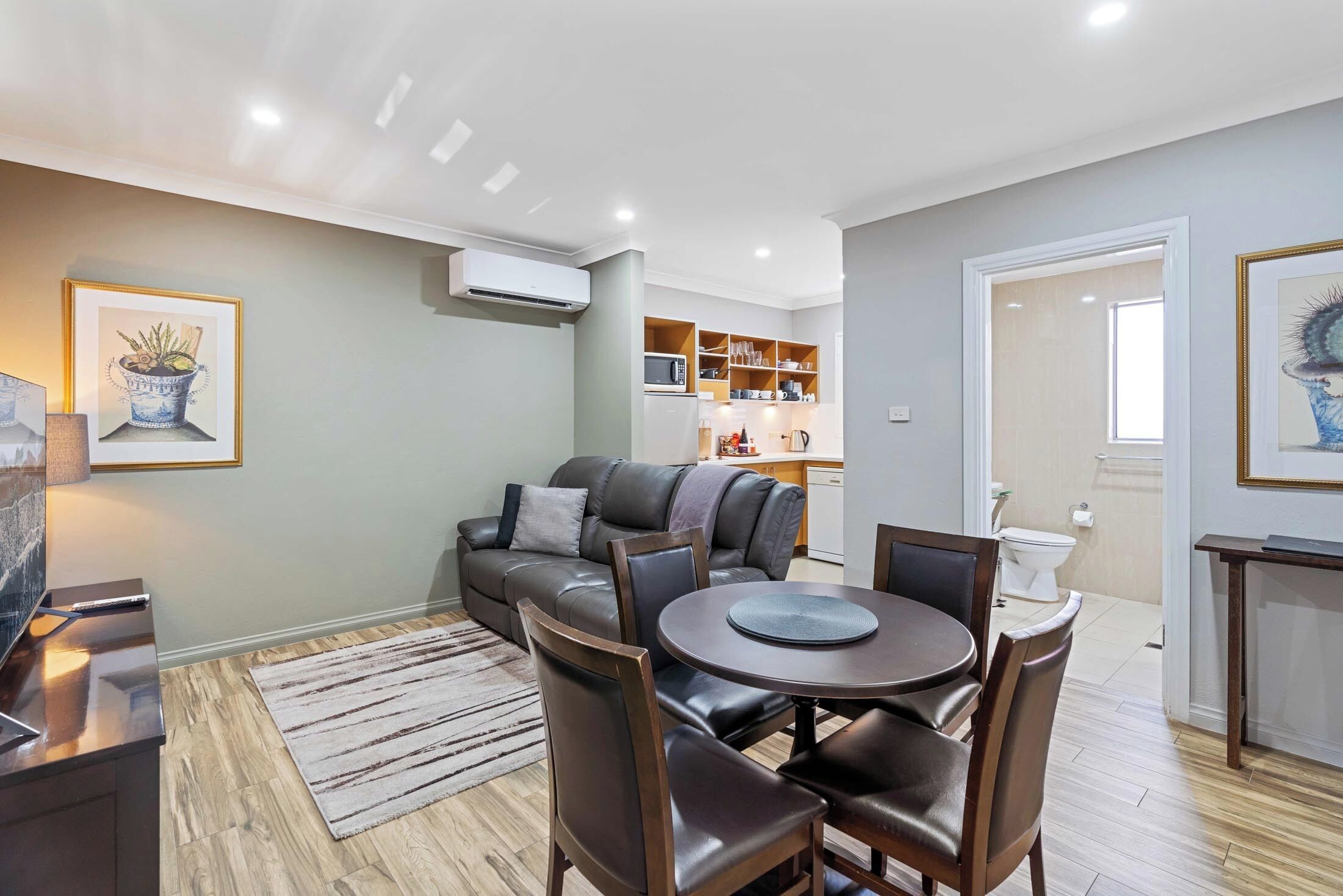 A modern living room with a leather sofa, dining table, and chairs. There is a kitchen area in the background with cabinets and a microwave.