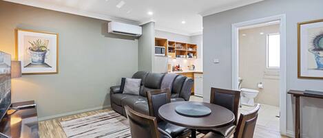 A modern living room with a leather sofa, dining table, and chairs. There is a kitchen area in the background with cabinets and a microwave.