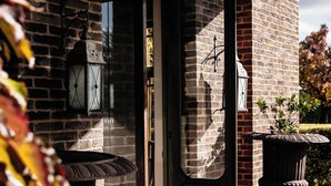 Fachada de edificio de ladrillo con puerta de madera, dos faroles en la pared y una planta en maceta.