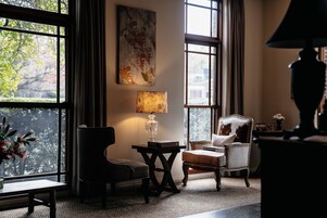 Una habitación acogedora con un amplio ventanal, una mesa con lámpara, una silla y un cuadro decorativo.