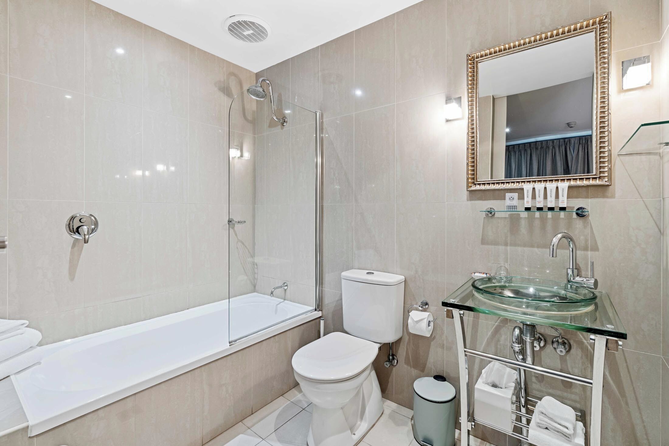 A modern bathroom with a glass-enclosed shower, a white toilet, a glass sink, and a mirror with a decorative frame.