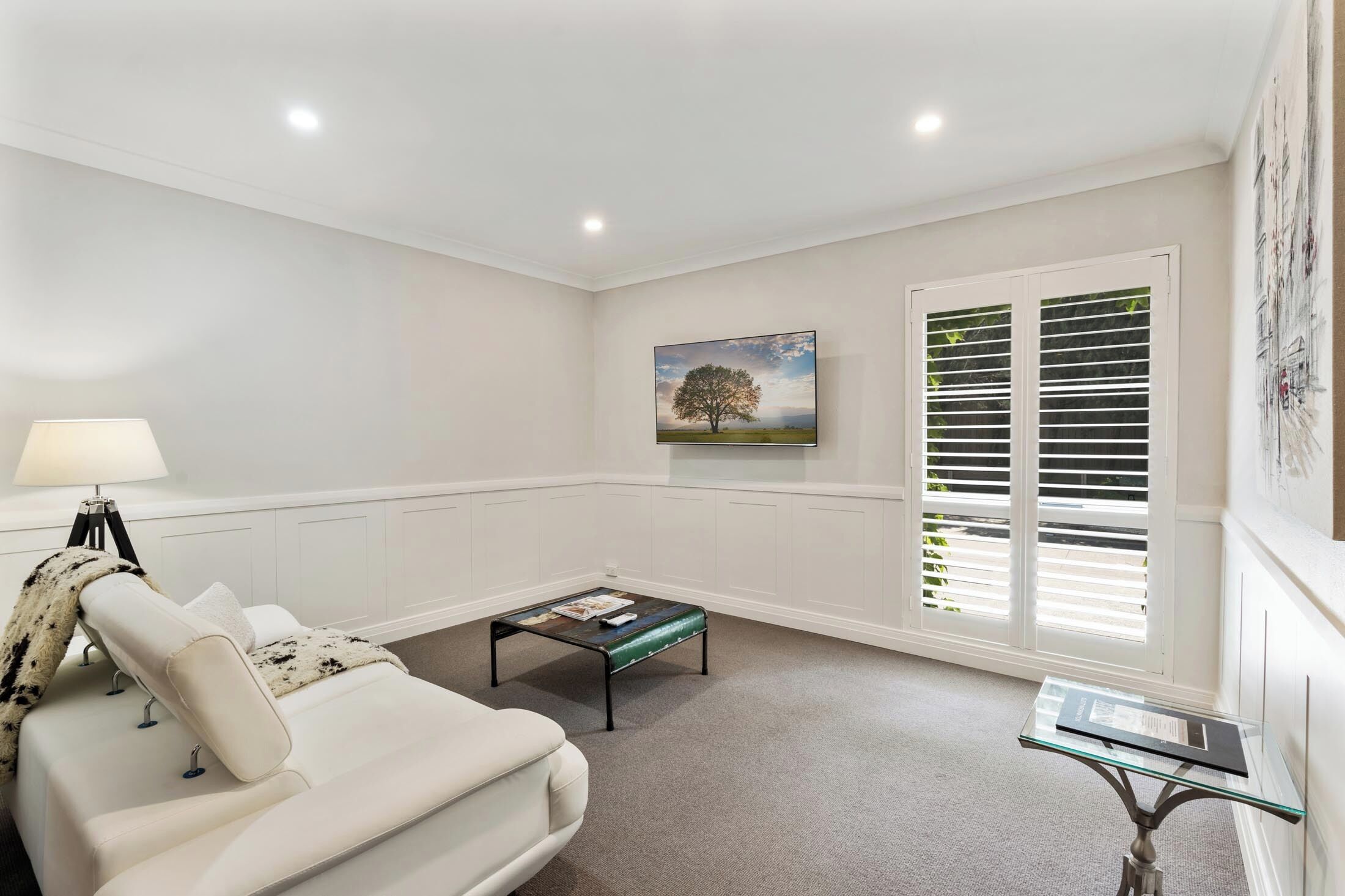 A modern living room with a white sofa, a glass coffee table, and a painting of a tree on the wall.