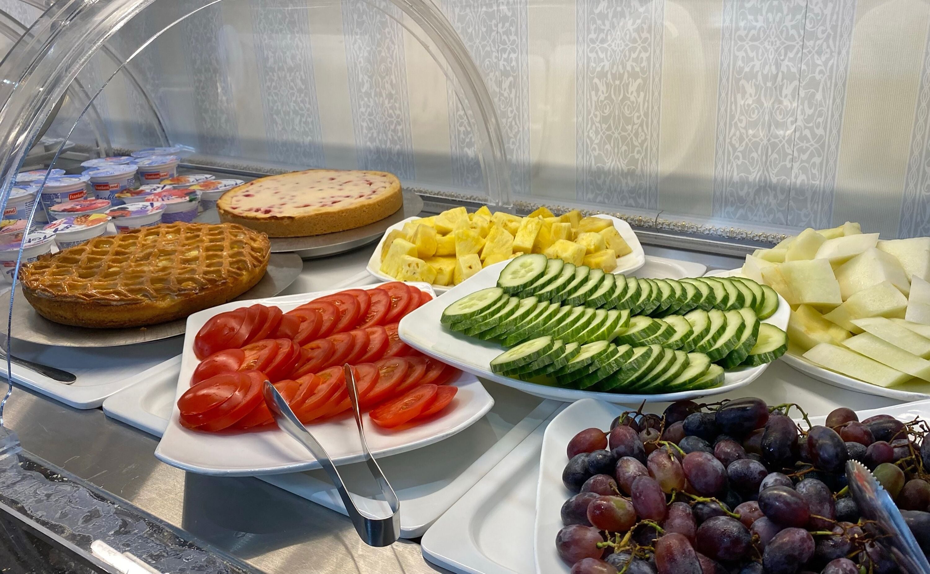 Une vitrine contenant divers aliments, notamment une tarte, des tomates en tranches, des concombres, des morceaux d’ananas et des raisins.