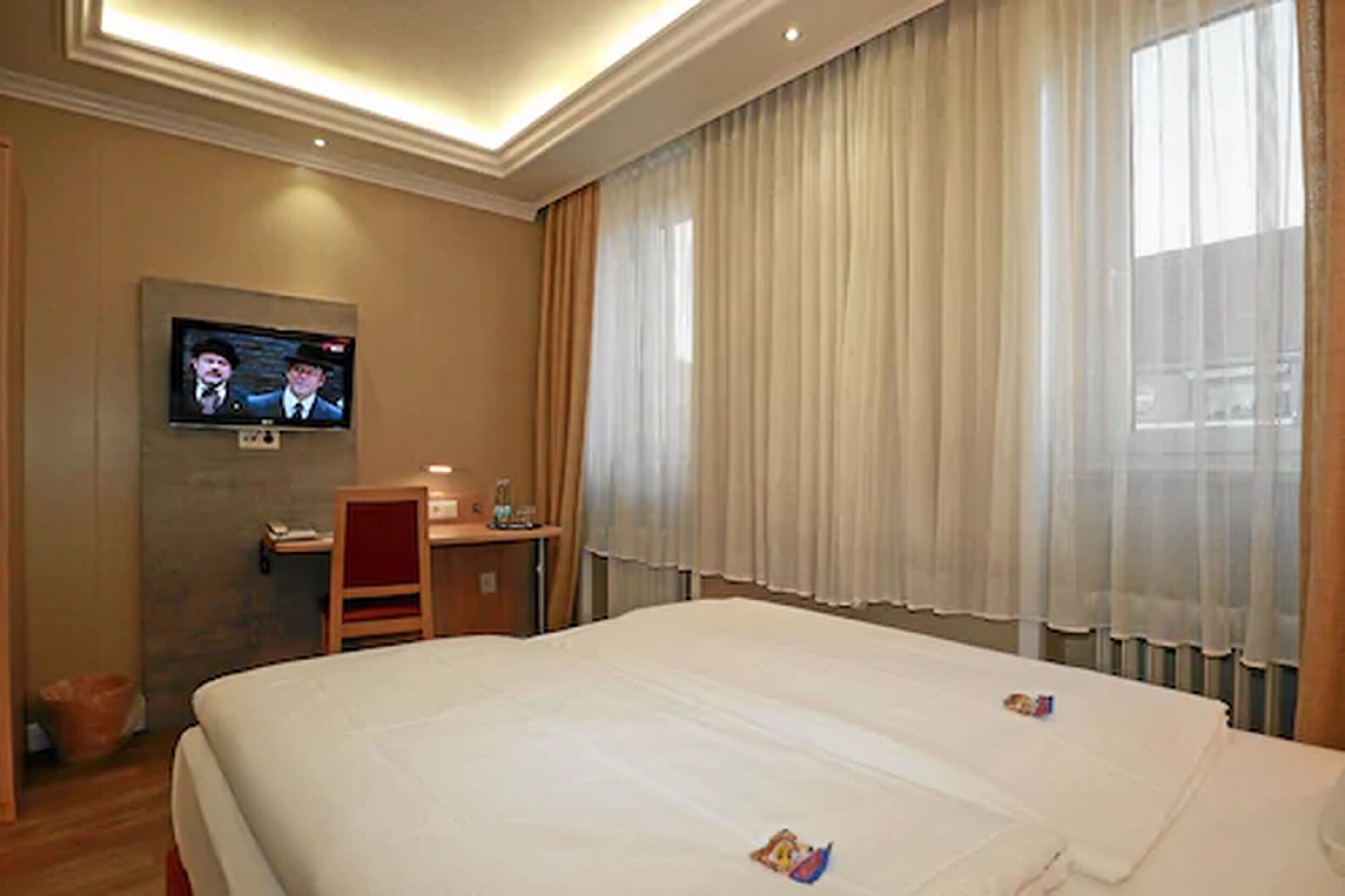 Ein Hotelzimmer mit einem großen Bett, einem Schreibtisch, einem Fernseher und einem Fenster mit Vorhängen.