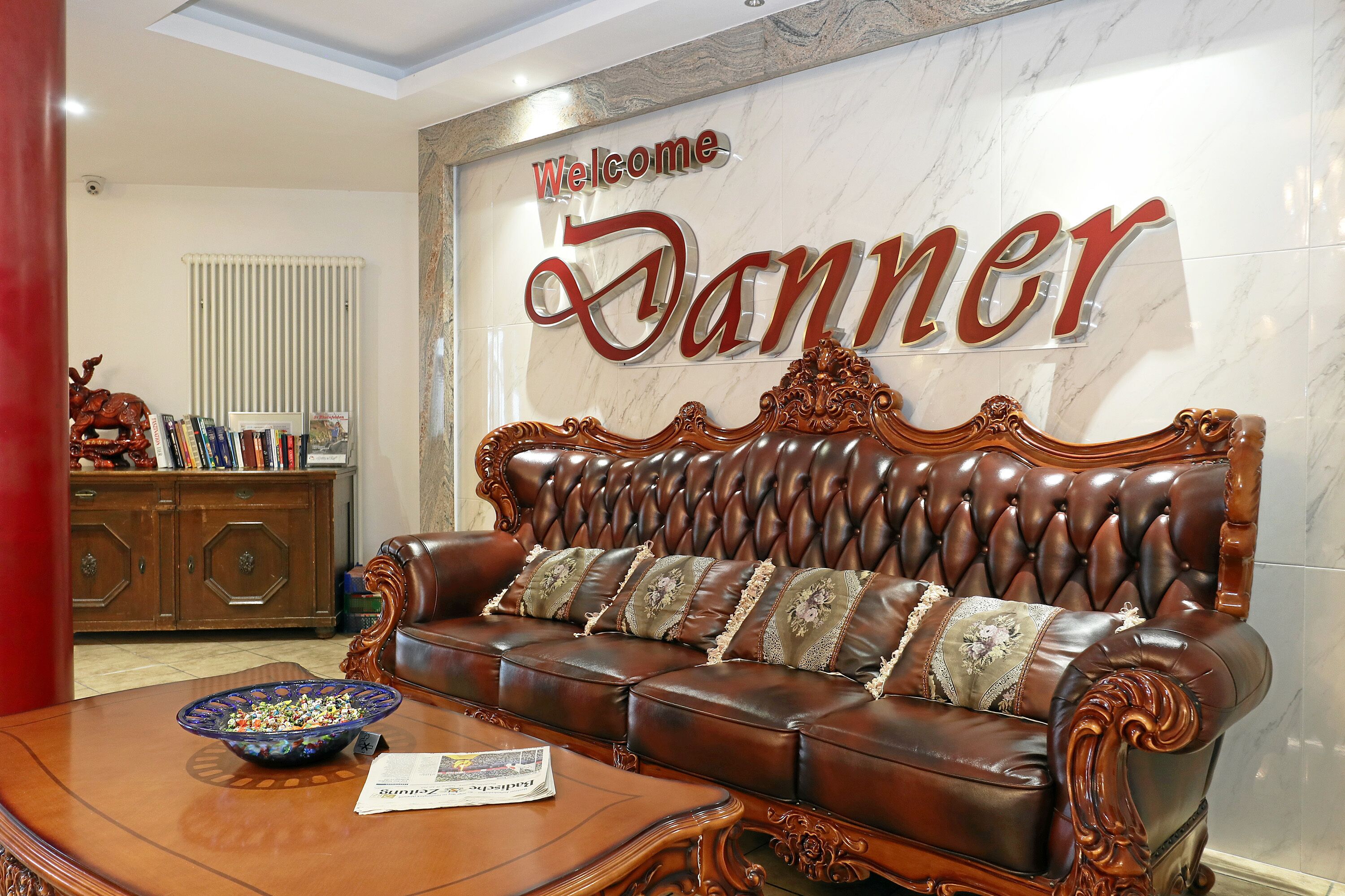 Een bruine leren bank met decoratieve kussens, een houten salontafel en een bordje met de tekst 'Welcome Danner' aan de muur.