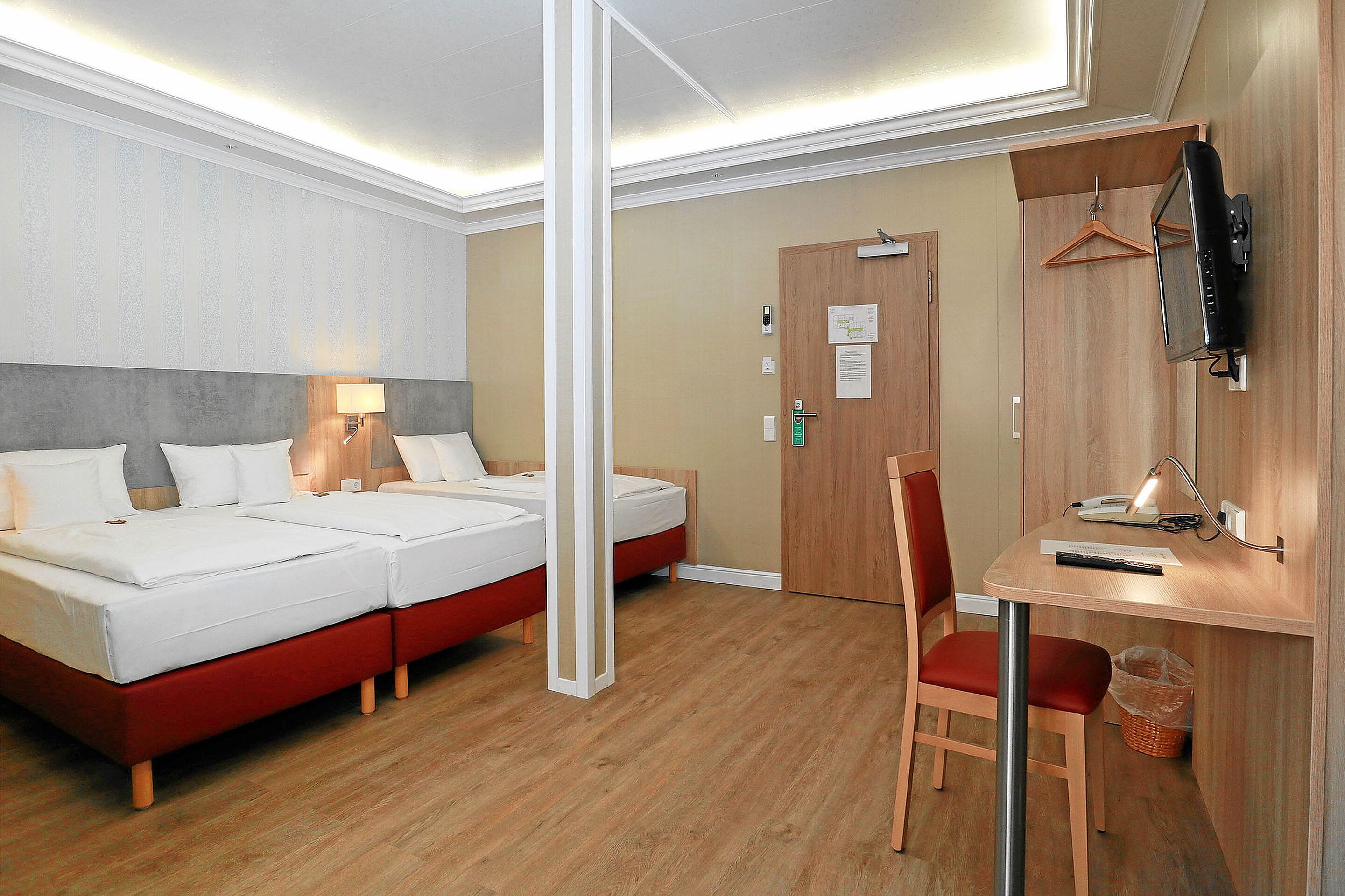 Habitación de hotel con cama doble, escritorio con silla, televisor montado en la pared y puerta de madera.