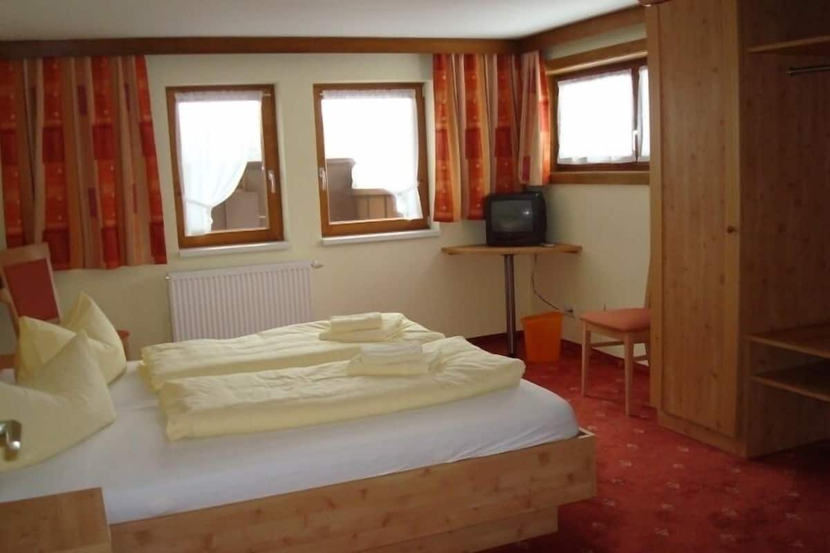 Une chambre avec un lit en bois, deux fenêtres avec des rideaux, un téléviseur posé sur une petite table et une chaise.