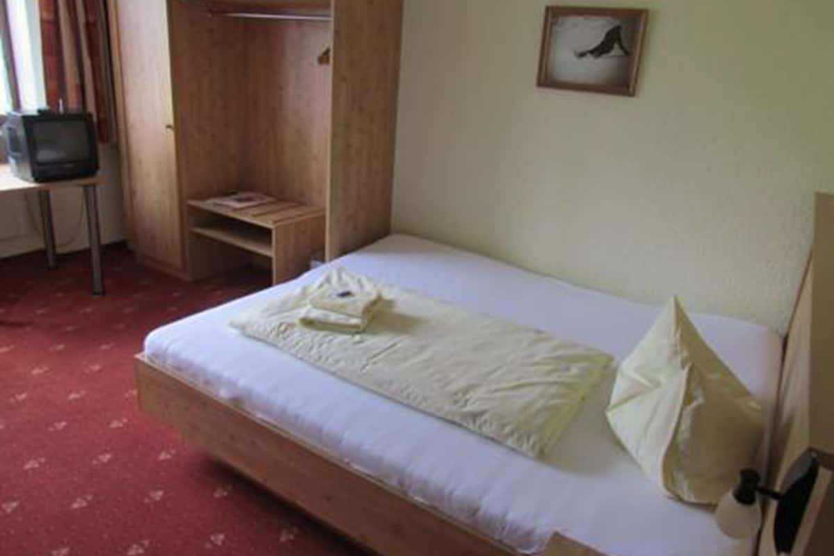 Une chambre avec un lit simple, un cadre de lit en bois, une petite table de chevet, un téléviseur posé sur un meuble et une armoire.