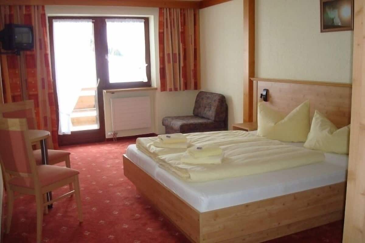 Une chambre d’hôtel avec un lit en bois, une chaise, un radiateur et une fenêtre avec des rideaux.