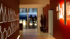 Un corridoio moderno di un hotel con un muro di bottiglie al neon, una porta in vetro che conduce a un balcone con vista sulla città e una reception con un parete rossa a contrasto.