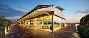 Un edificio moderno con techo inclinado y grandes ventanales, con terraza al aire libre y vistas al agua.