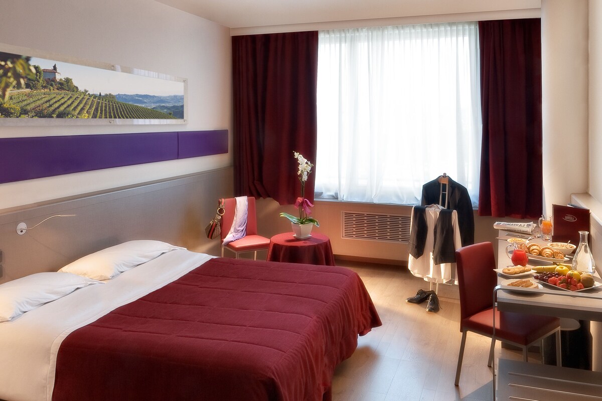 A hotel room with a bed, a desk with breakfast, a chair, a window with curtains, and a painting on the wall.
