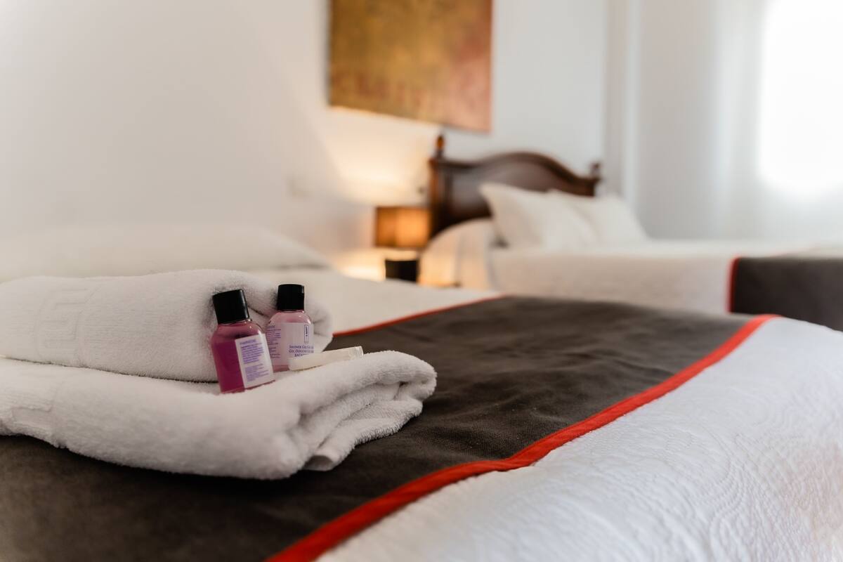 Ein ordentlich bezogenes Bett mit weißer und roter Tagesdecke, ein gefaltetes weißes Handtuch und zwei kleine Lotionflaschen.