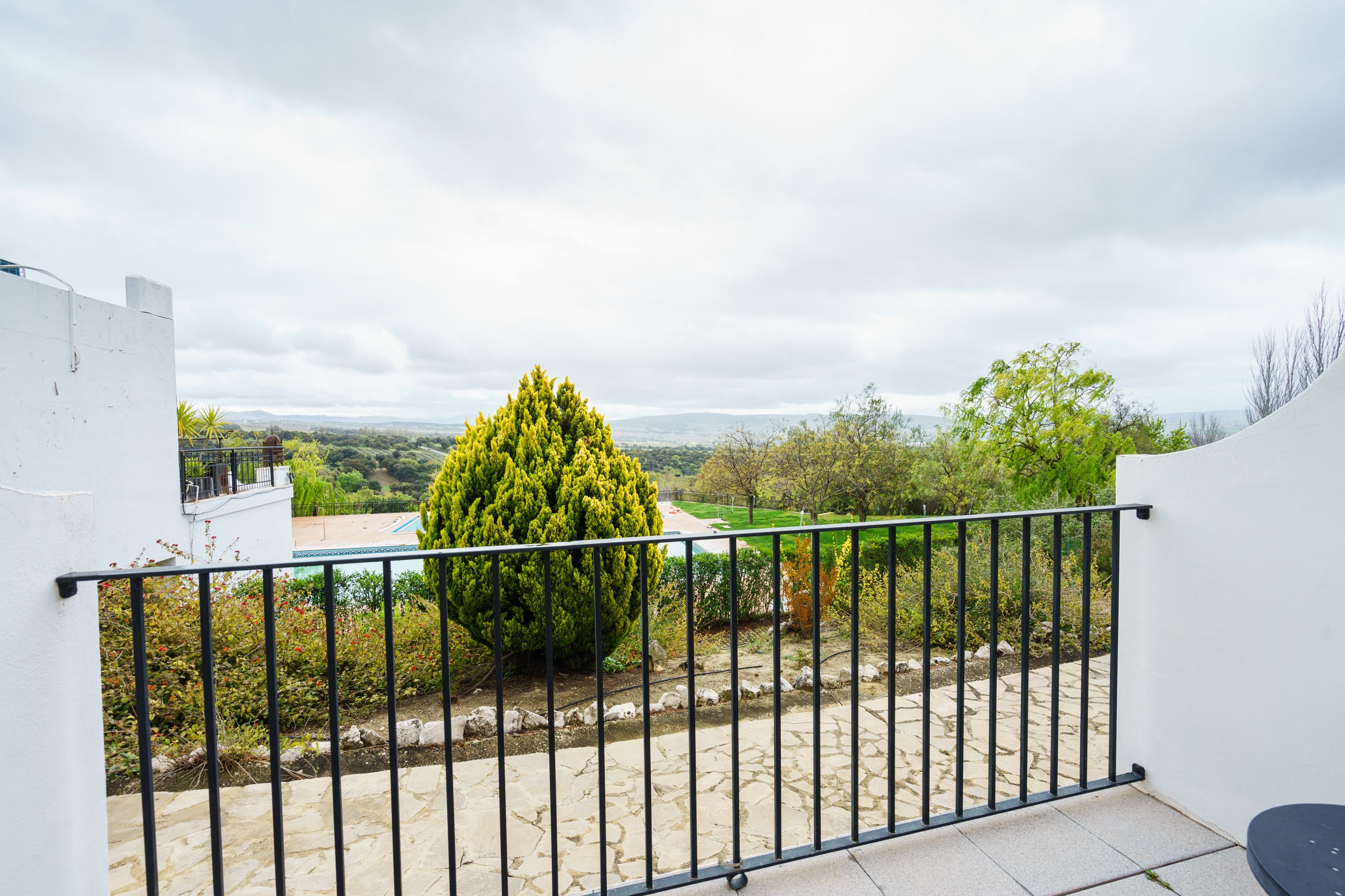 Ein Balkon mit Steinboden, einem Metallgeländer und Blick auf eine grüne Landschaft mit Bäumen und einem entfernten Gebirge.