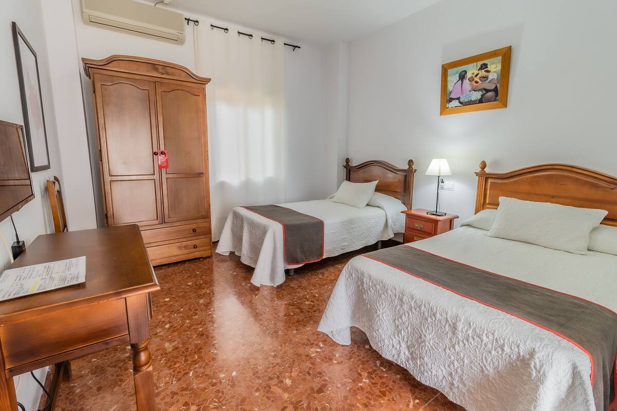 Ein Hotelzimmer mit zwei Betten, einem Holzschrank, einem Schreibtisch mit Stuhl, einem Bild an der Wand und einem Fenster mit Vorhängen.