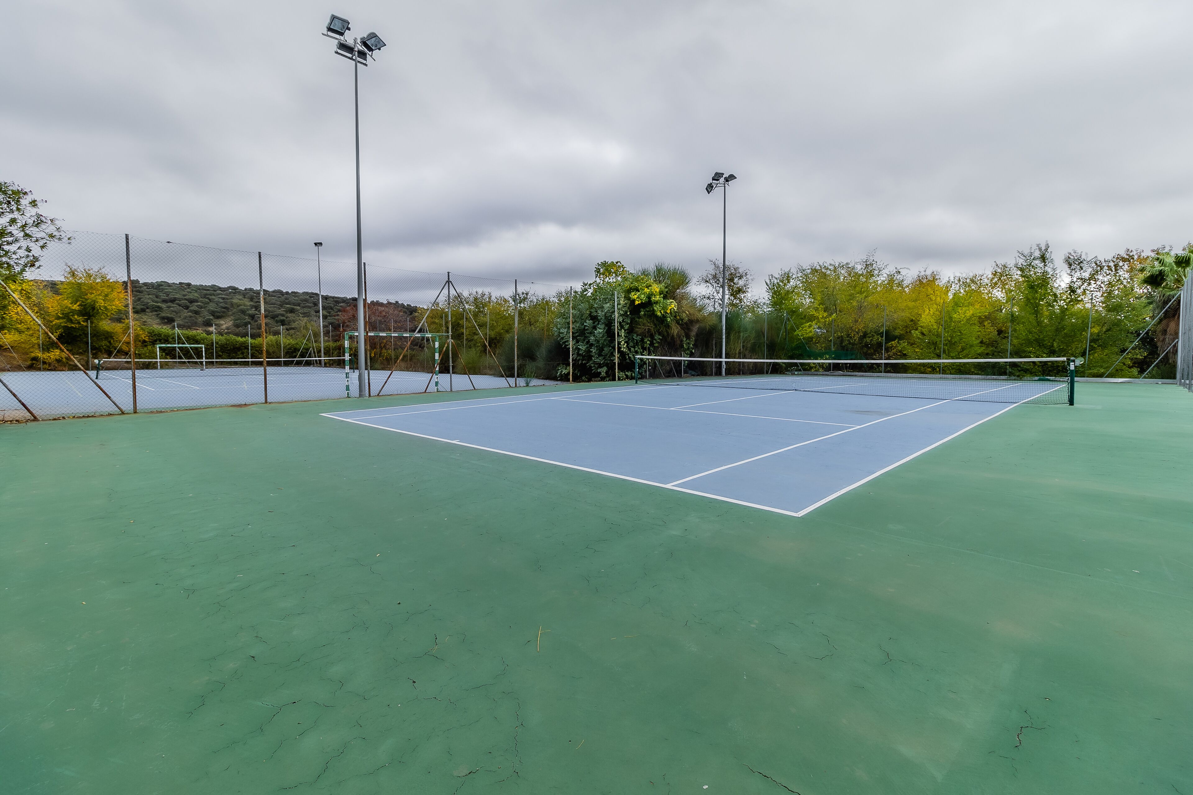 An outdoor tennis court with a net, surrounded by a fence, and under floodlights.