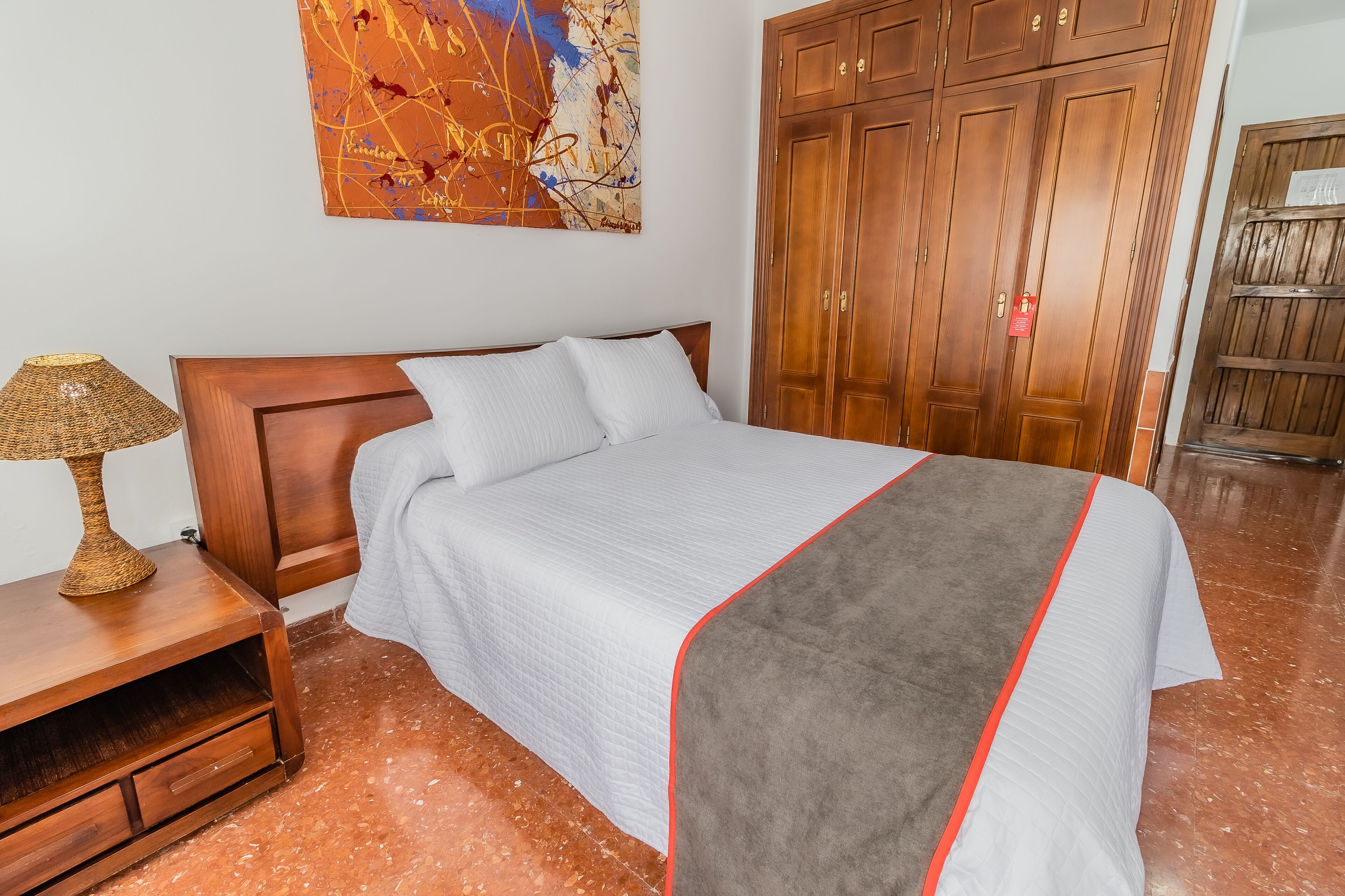 A neatly made bed with a white and grey blanket, a wooden nightstand with a lamp, and a large wooden wardrobe.