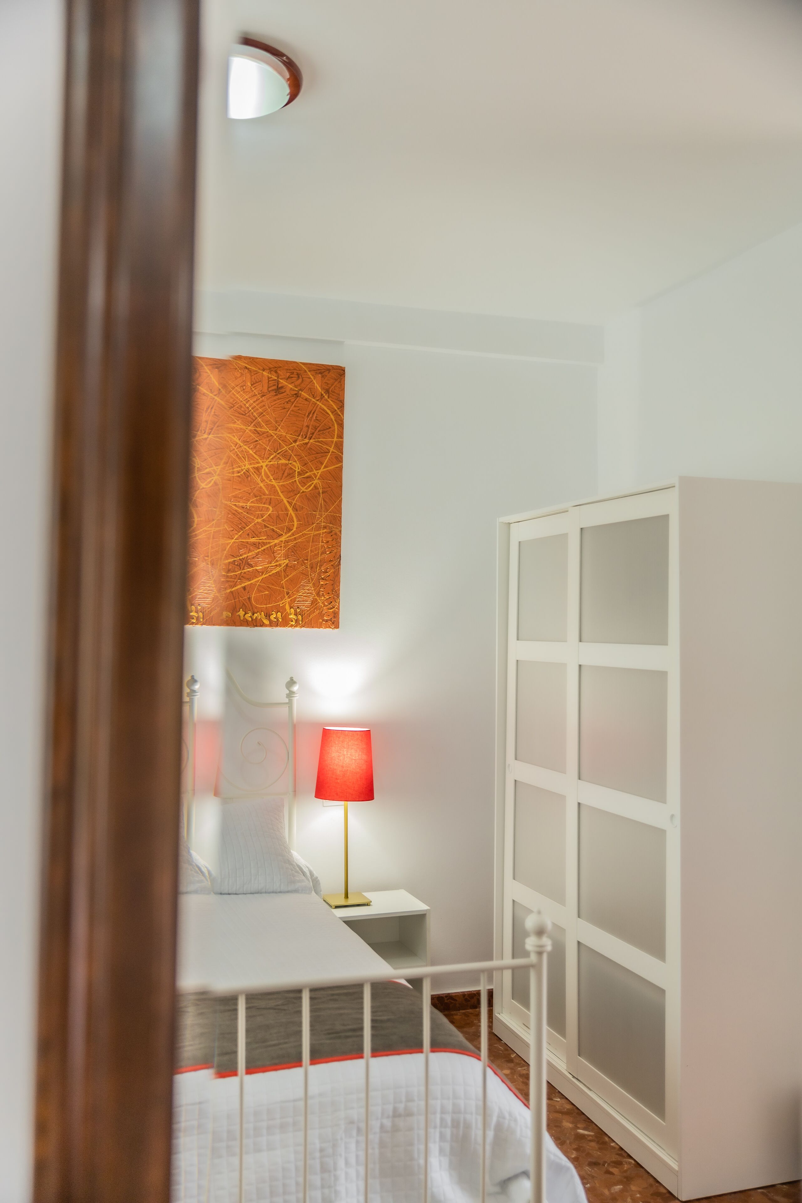 Quarto com cama, mesa de cabeceira, abajur vermelho, guarda-roupa branco e uma parede com uma pintura abstrata.