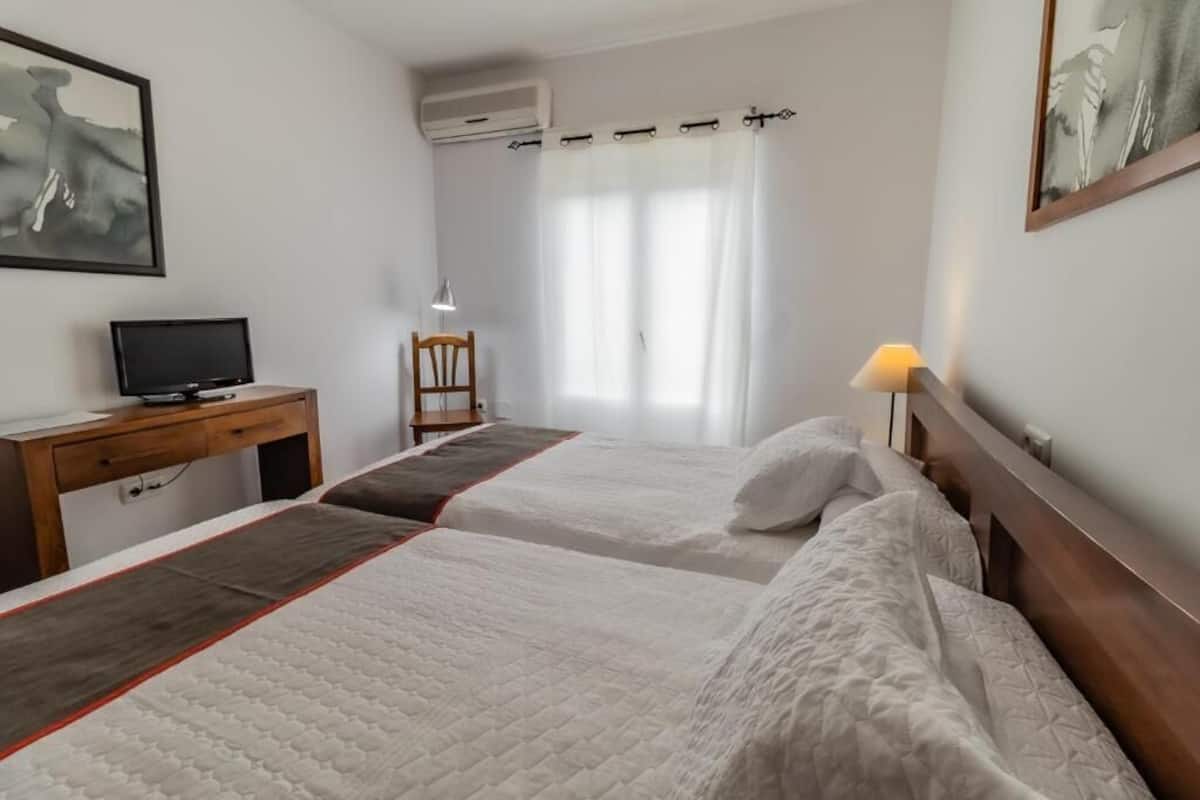 Ein Hotelzimmer mit zwei Betten, einem Schreibtisch mit Computer, einem Stuhl, einer Lampe und einem Fenster mit Vorhängen.