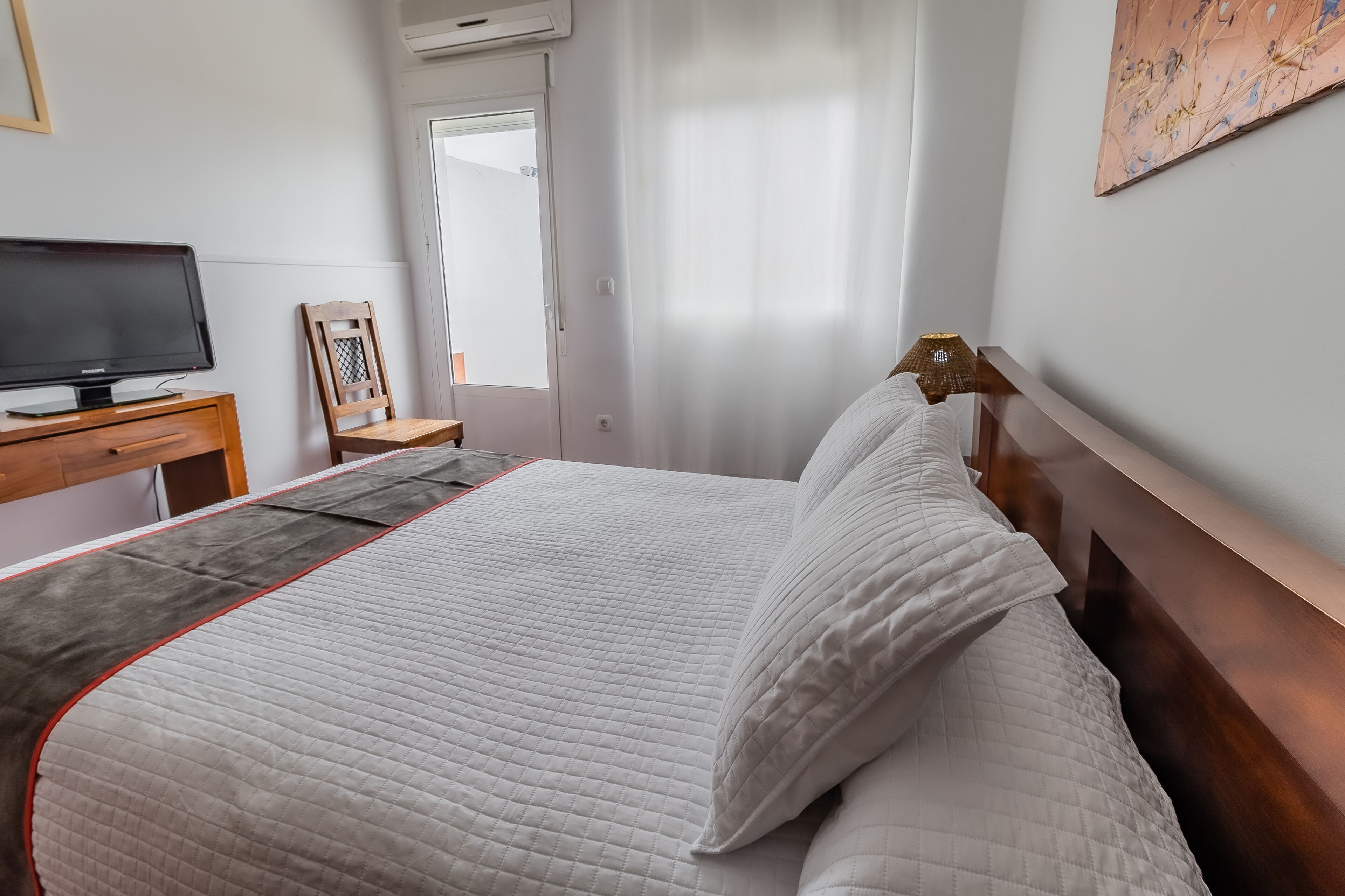 A neatly made bed with a quilted cover, a wooden bedside table with a flat-screen TV, a chair, and a window with curtains.