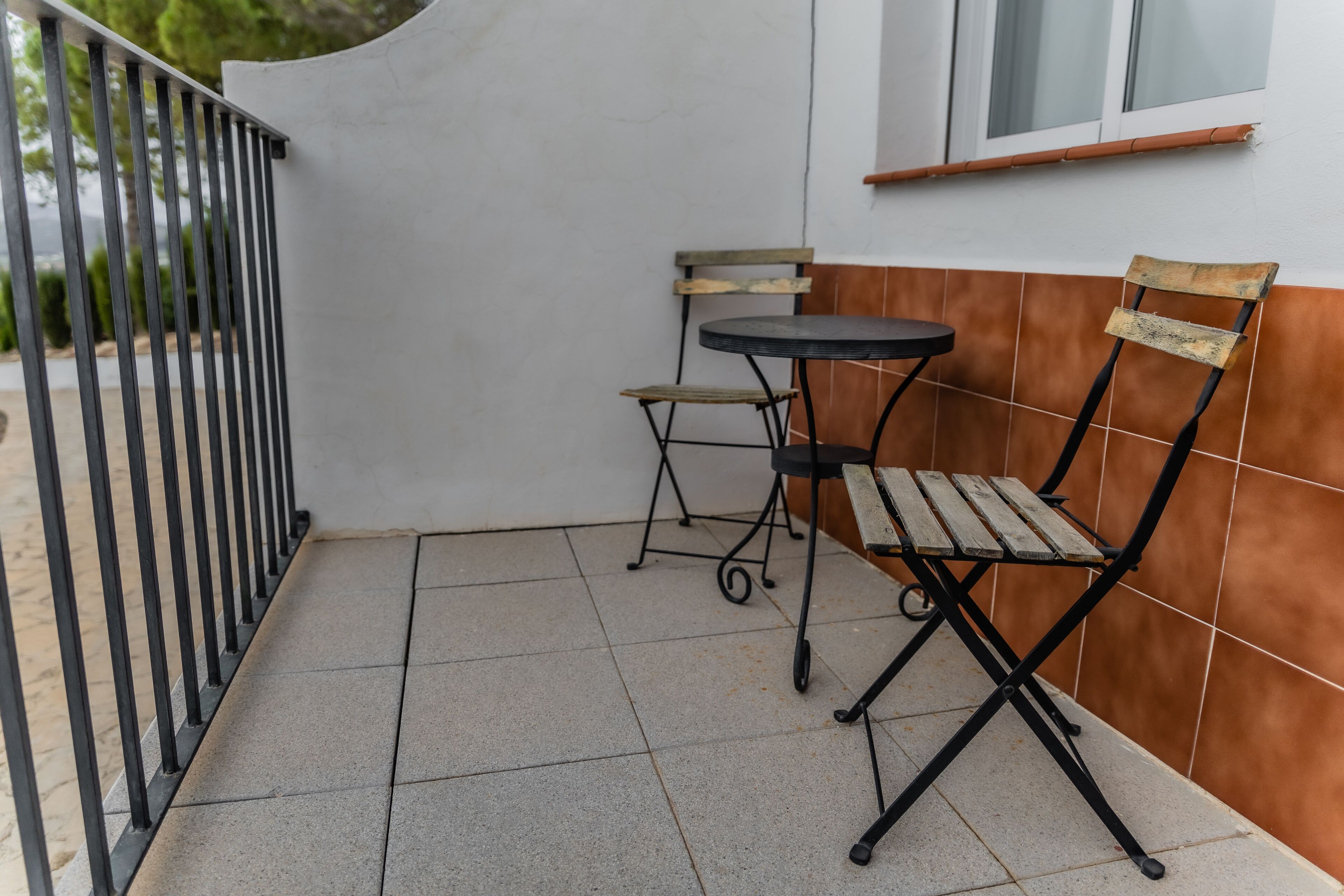 A small outdoor patio with a metal railing, a foldable table, a foldable chair, and a window with blinds.