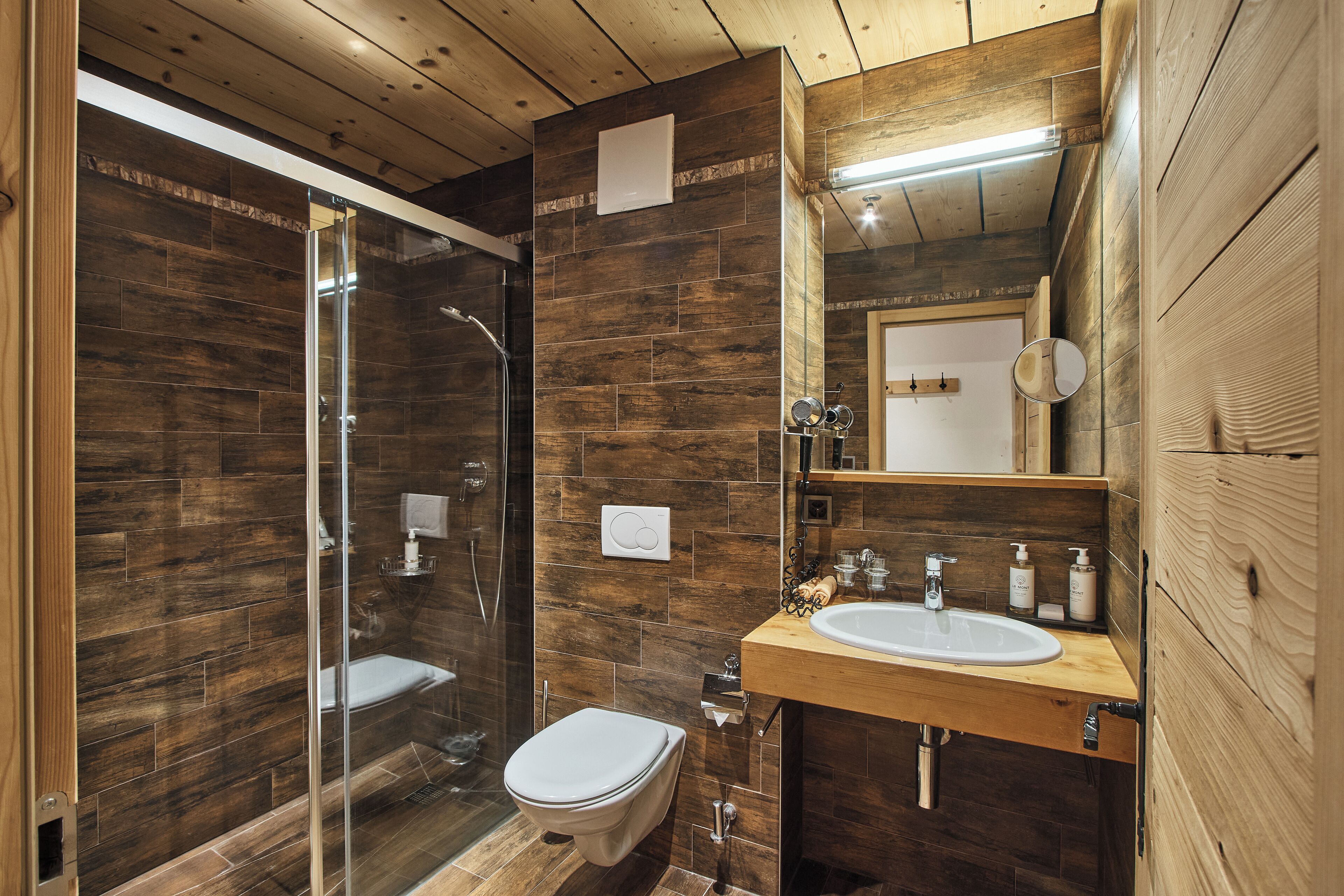 Un baño moderno con suelo de madera, cabina de ducha de vidrio, inodoro blanco y tocador de madera con lavamanos y espejo.