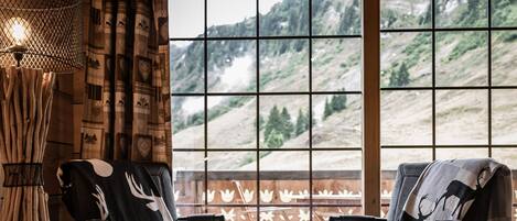 A room with a bed, two chairs, a small table, and a view of a mountainous landscape through large windows.