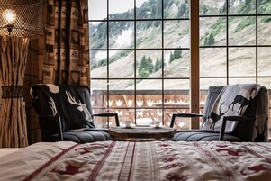 Una habitación con una cama, dos sillas, una mesita y vistas a un paisaje montañoso a través de amplias ventanas.