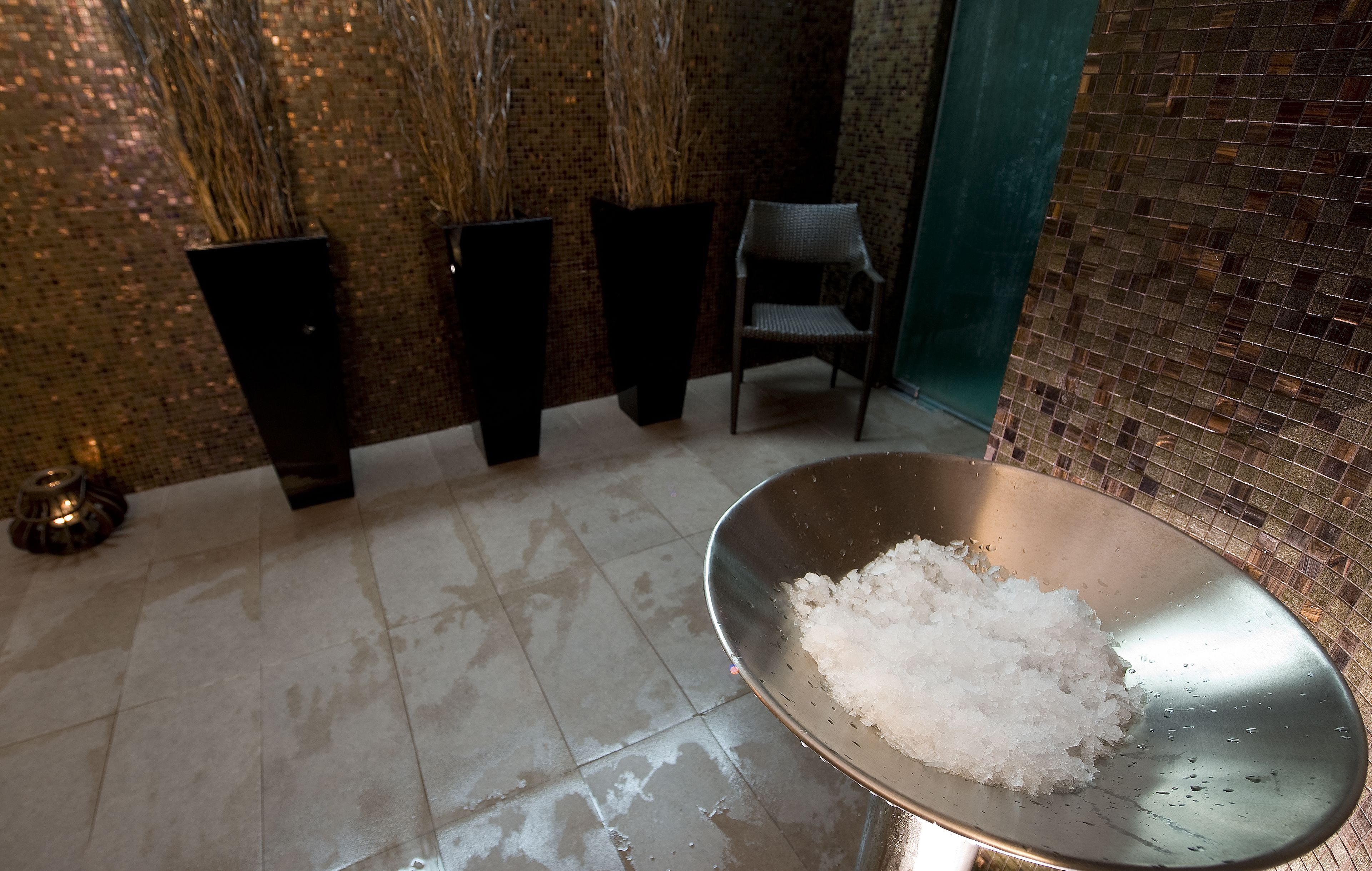 A room with a tiled floor, a metal bowl with salt, and three tall black planters with dried plants.