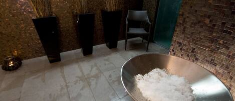 A room with a tiled floor, a metal bowl with salt, and three tall black planters with dried plants.