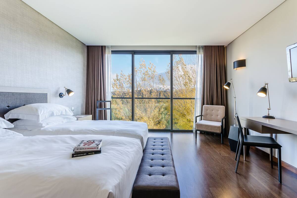 A hotel room with two beds, a desk, a chair, and a large window with a view of trees.