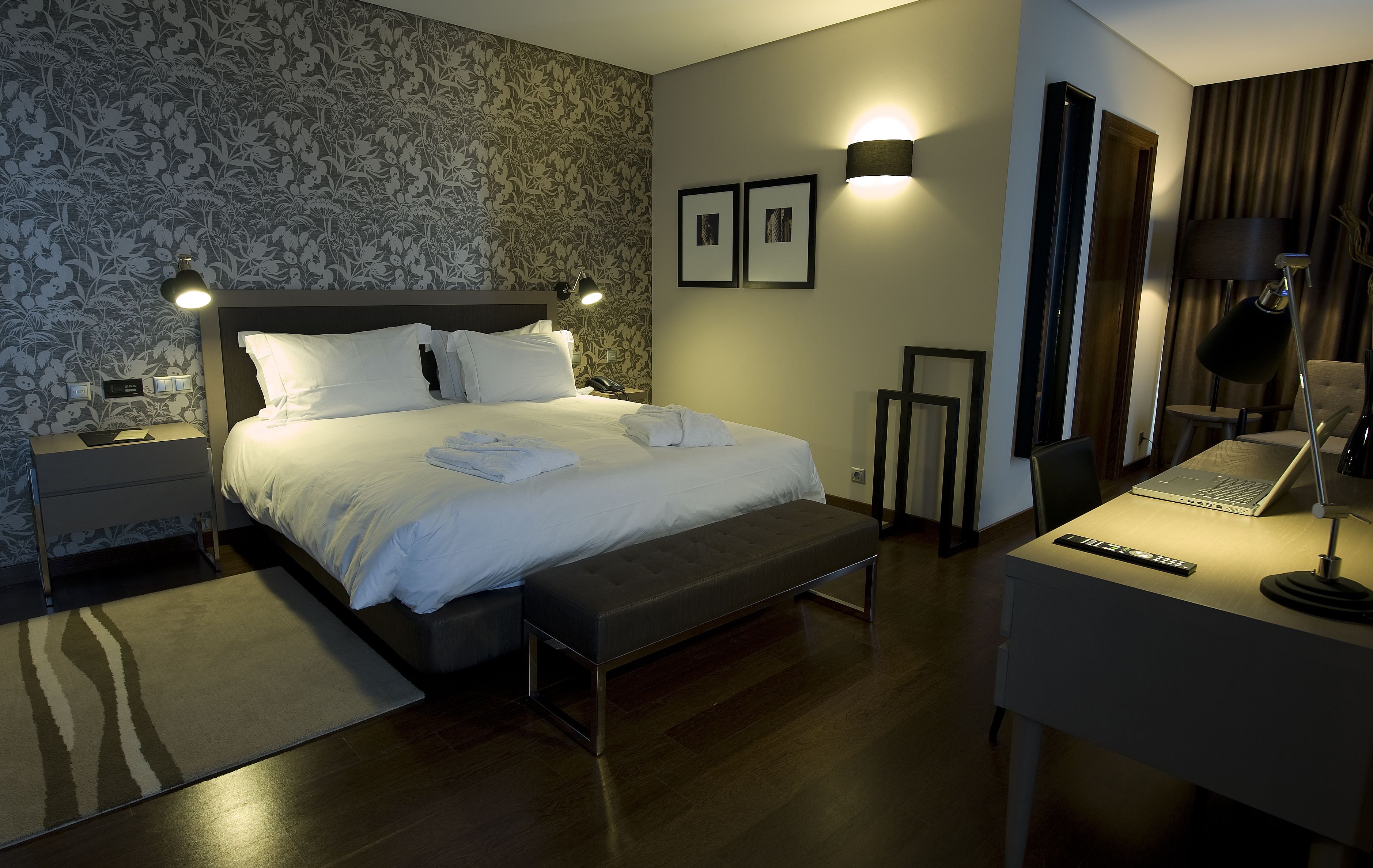 A modern hotel room with a large bed, bedside lamps, a desk with a laptop, and a chair.