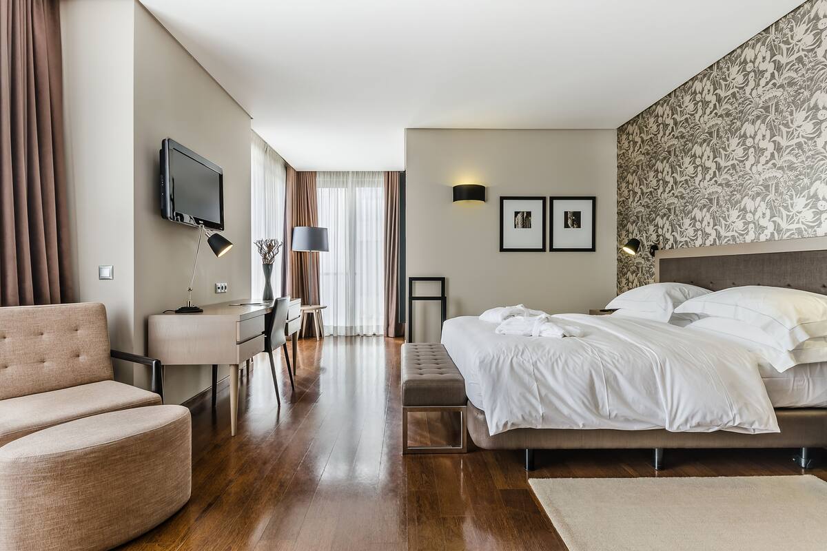 A modern hotel room with a bed, a desk, a chair, a TV, and a large window with curtains.