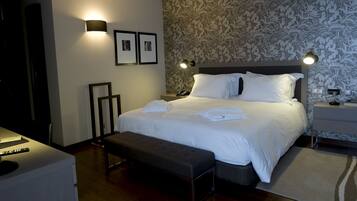 Una cama bien tendida con sábanas blancas, una mesita de noche con lámpara y un banco en una habitación de hotel con papel tapiz estampado.