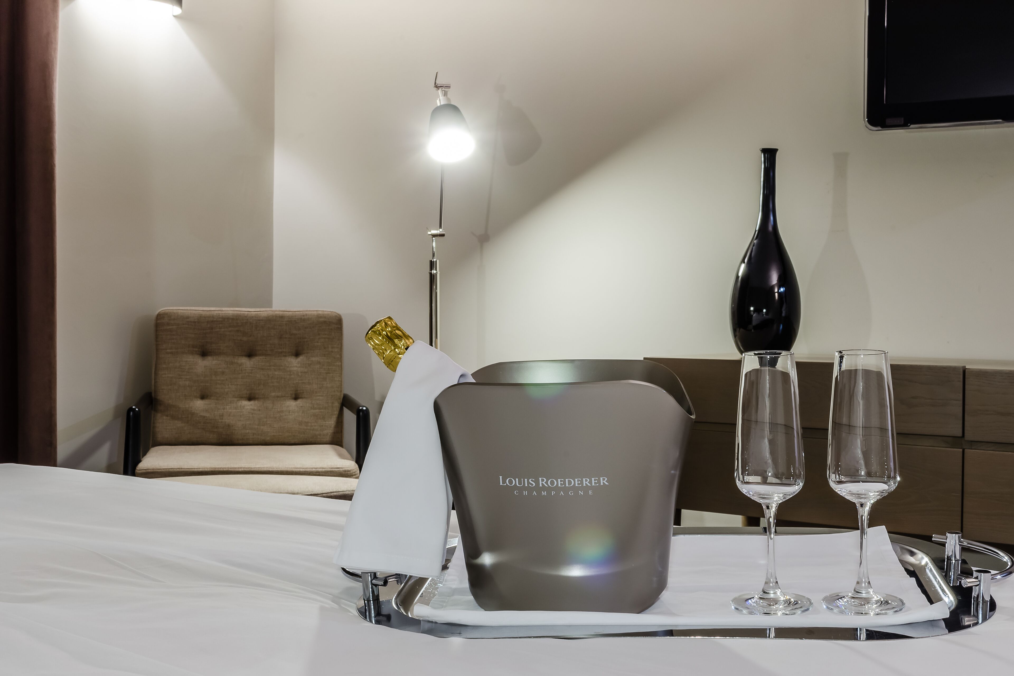 A hotel room with a bed, a bedside table with a bottle of champagne, two glasses, and a tray.