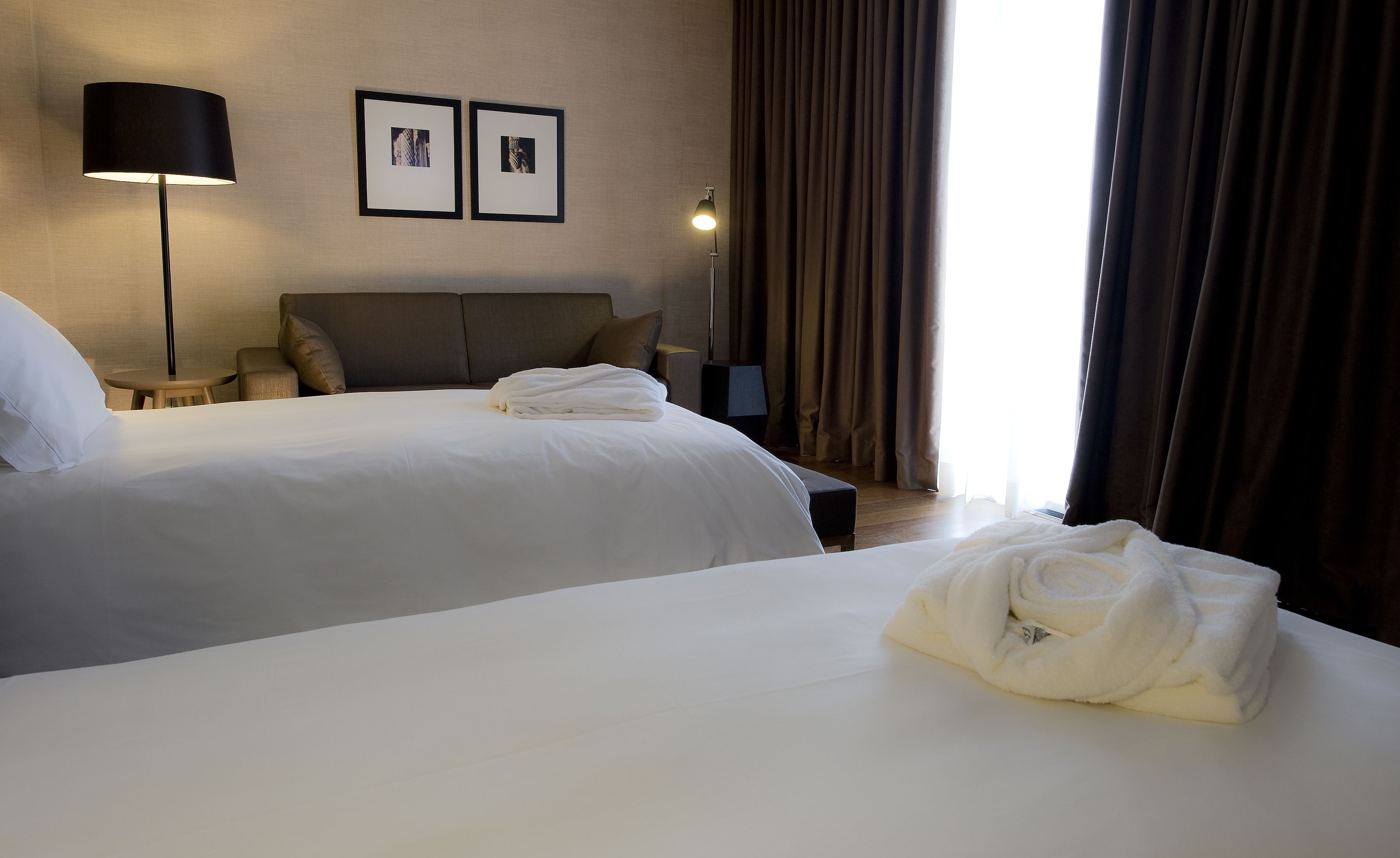 A hotel room with two beds, a lamp, a small table, and a window with curtains.