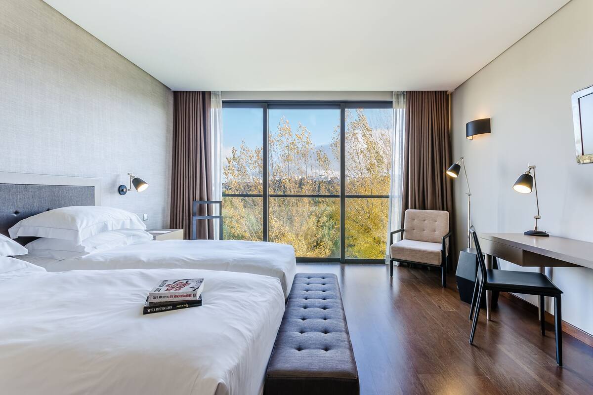 A modern hotel room with two beds, a desk, a chair, and a large window with a view of trees.