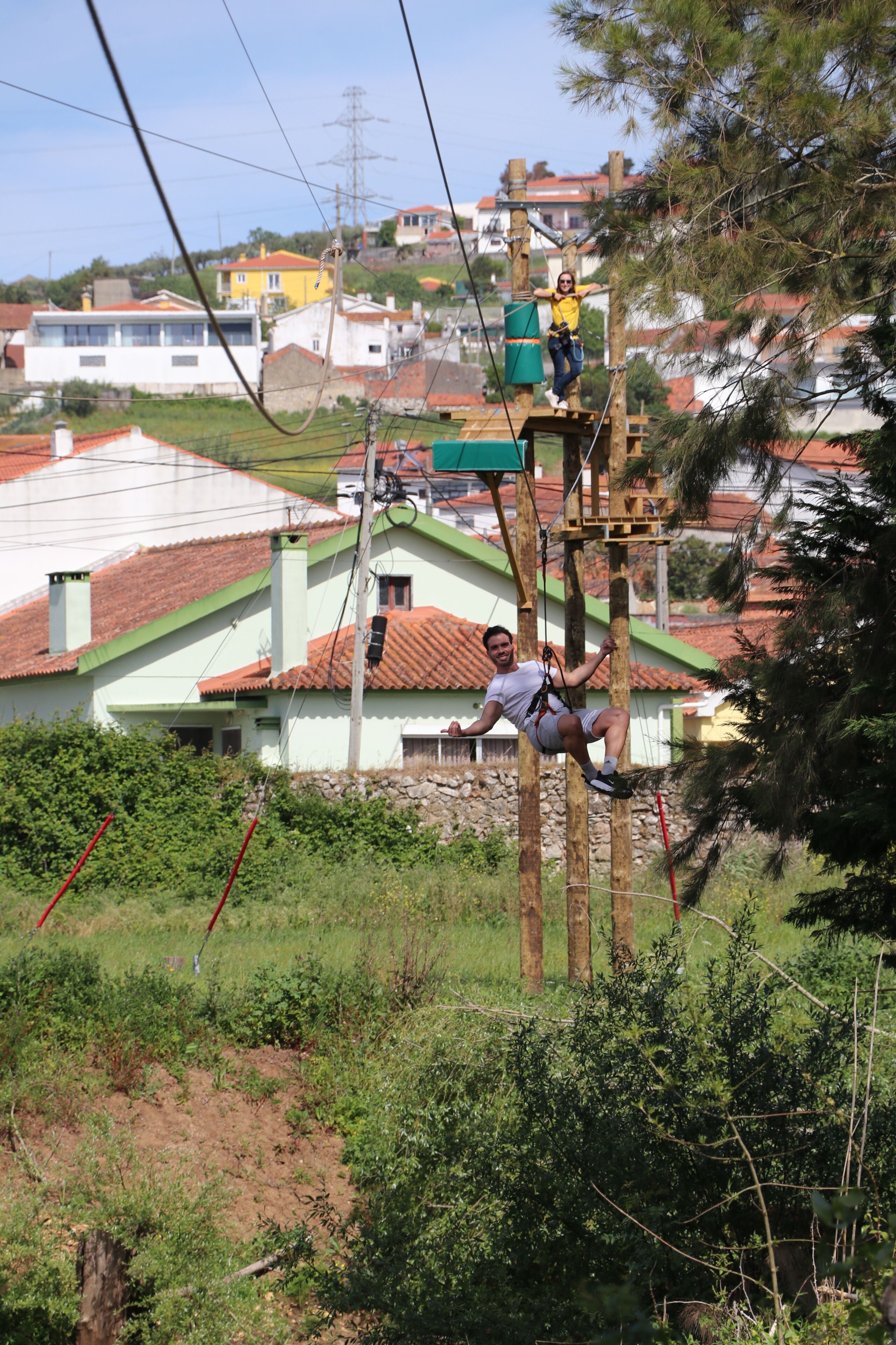 A person zip-lining over a wooden structure with a rope, with houses and greenery in the background.