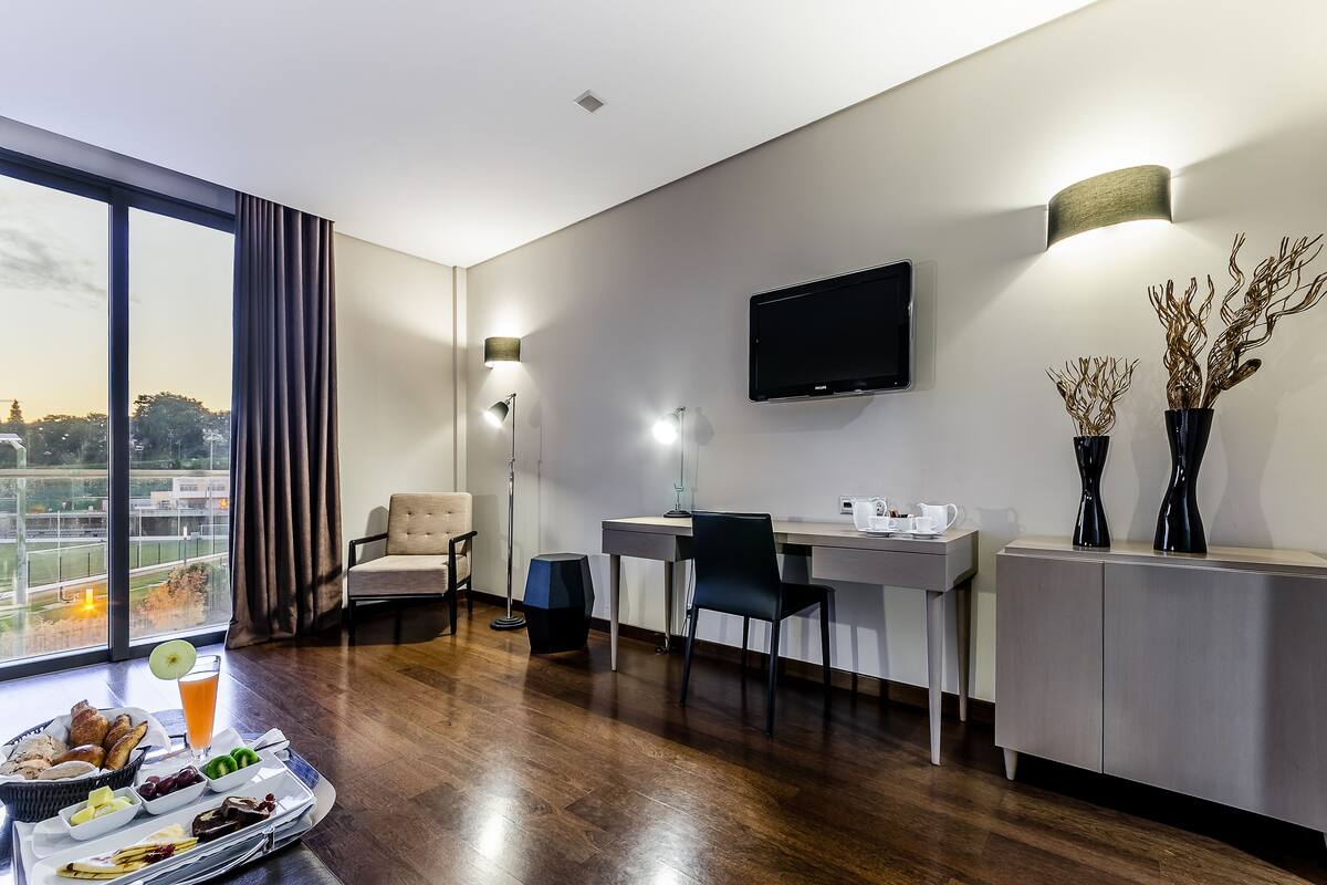 A modern hotel room with a large window, a desk with a chair, a flat-screen TV, and a dining area with a tray of food.