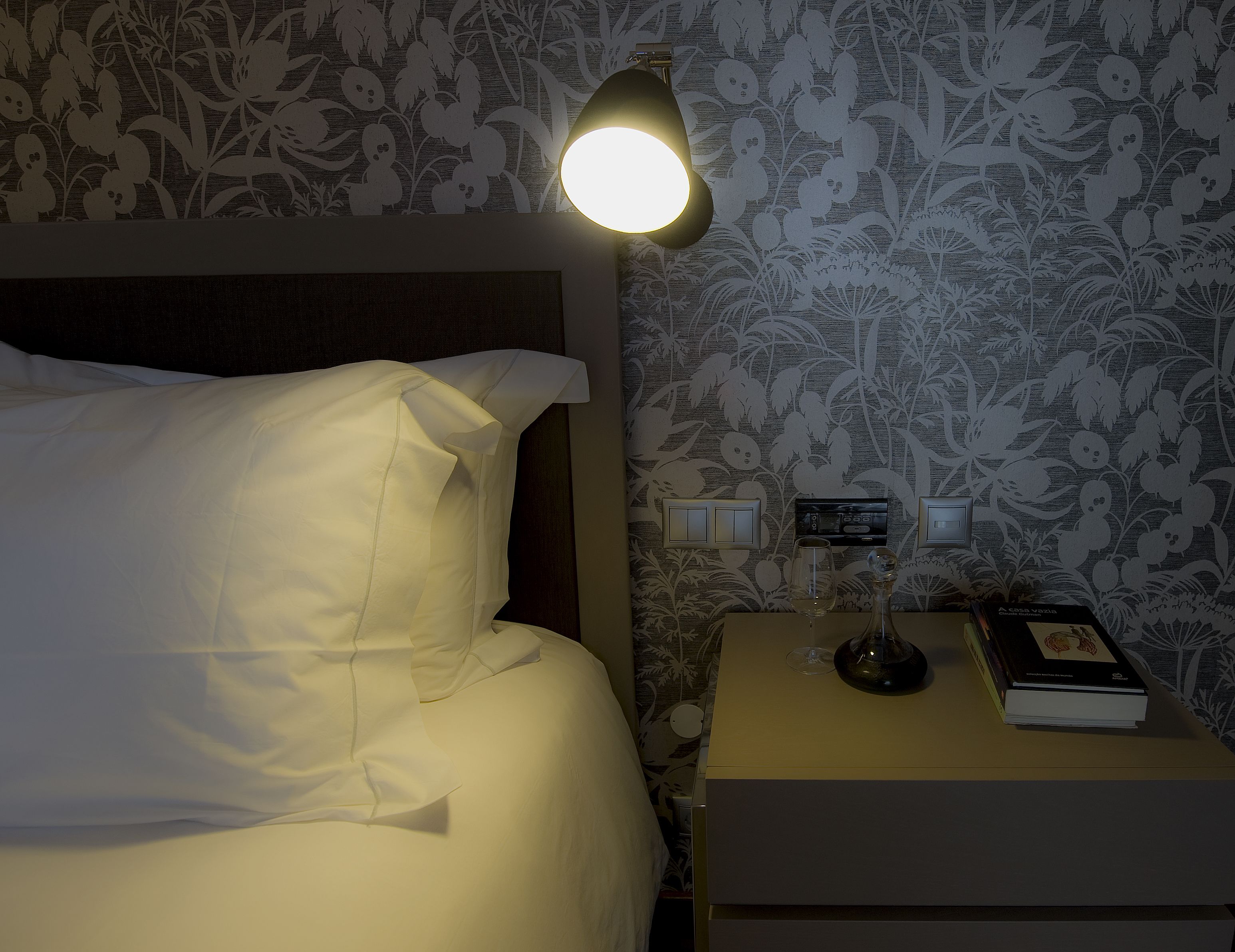 A neatly made bed with white linens, a bedside lamp, a nightstand with a book and a glass, and a wall with floral wallpaper.