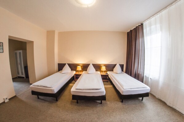 A hotel room with three single beds, each with a bedside table and lamp, a large window with curtains, and a door leading to another room.