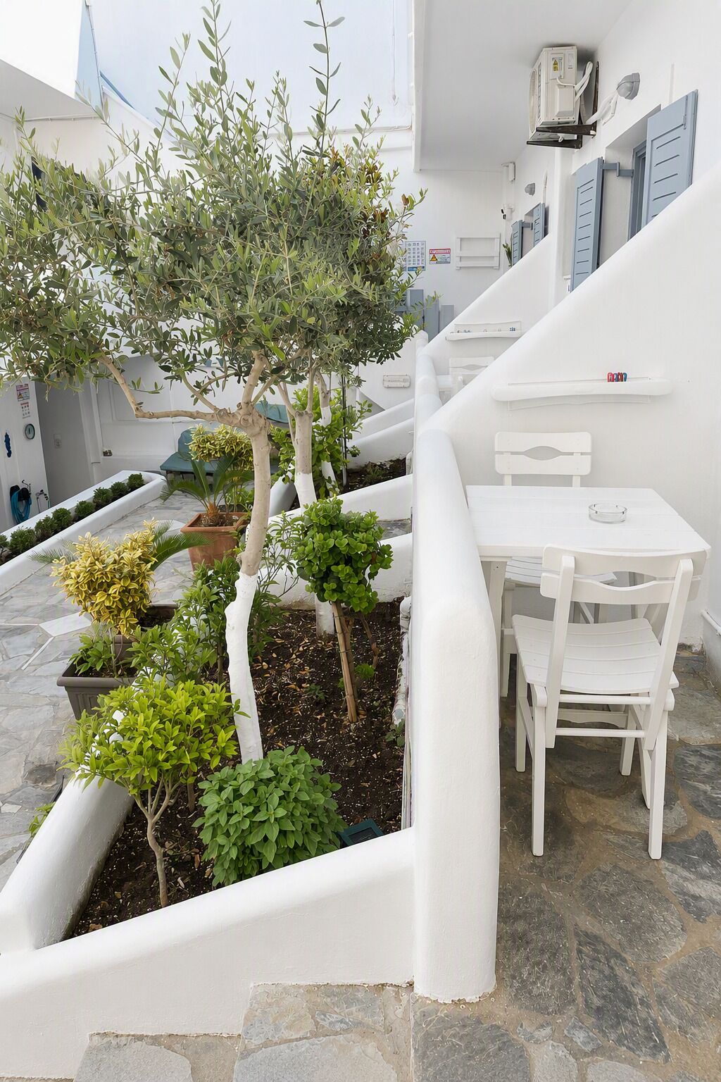 A white outdoor seating area with a table and chairs, surrounded by potted plants and a small tree.